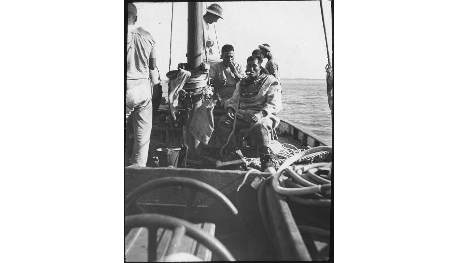 Black and white photo of Japanese men on a pearl diving boat, one dressed in diving attire but with the helmet off