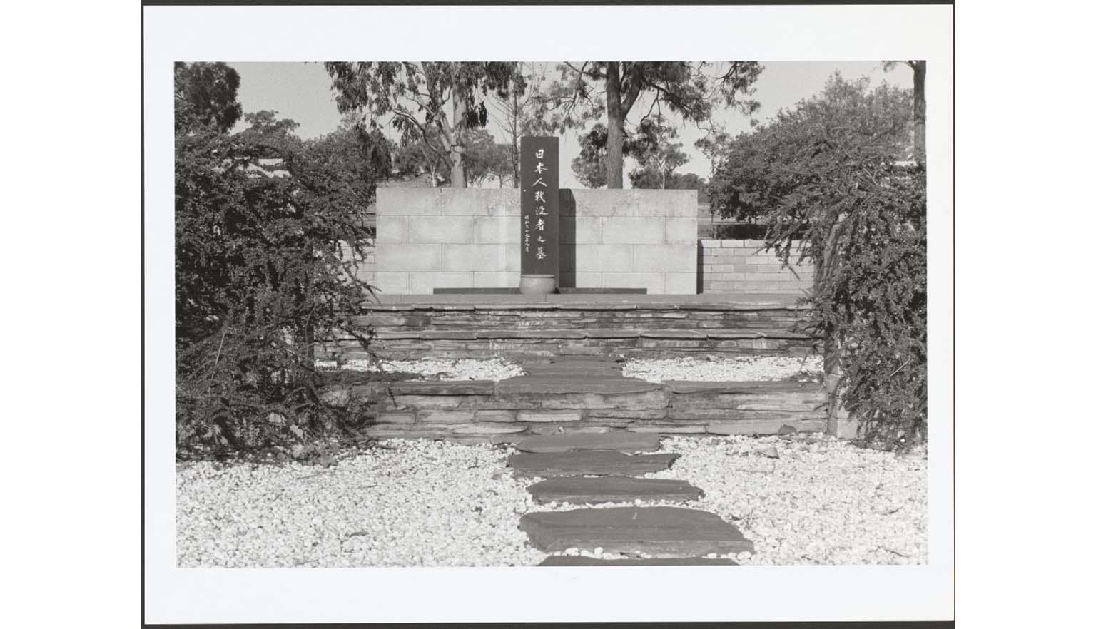 A black and white photograph of a stone memorial. In the middle of the memorial is a black stone obelisk with carved Japanese kanji. The site is surrounded by trees. There is a flagstone path surrounded by white pebbles leading up to the memorial