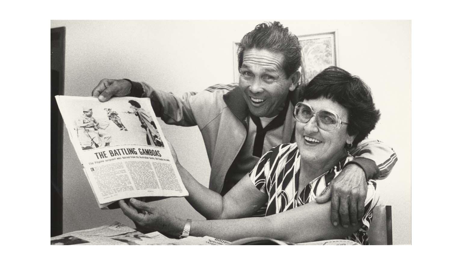 usband and wife holding up a newspaper article about themselves, with their arms around each other and large smiles