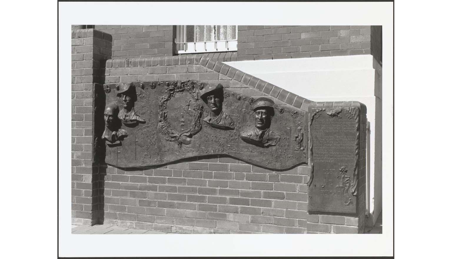 A black and white photograph of a memorial plaque. The plaque is attached to a brick wall in front of a building. The plaque features bas relief sculptures of the busts of 4 men. Two are wearing AIF slouch hats, another wears an officers flat cap and the other has no had. There is a rectangular section to the right with the words 'Lest we forget'
