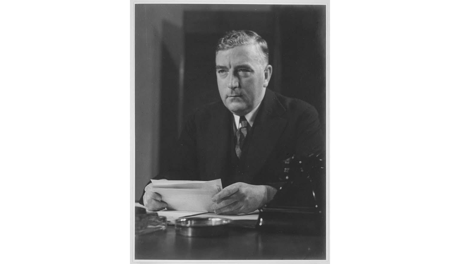 Australian Prime Minister Robert Menzies sits at a desk holding several pieces of paper. He is looking melancholic and stares off camera to the left. He is a middle-aged white man with blonde hair swept to one side. He wears a dark suit and tie with a dark tie. There is a black Bakelite telephone on the table in front of him