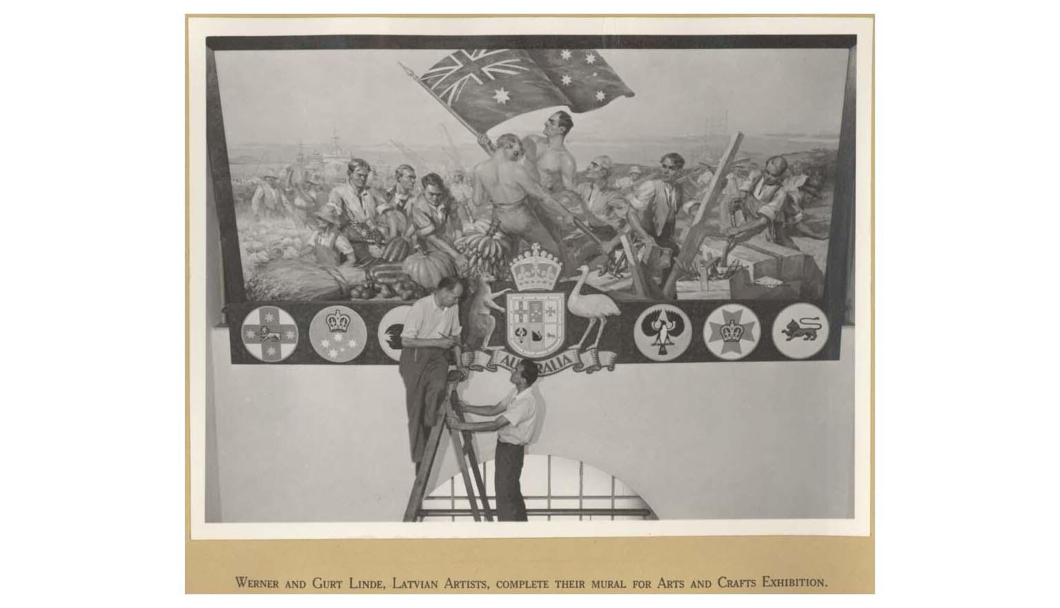 Two men, one standing on a ladder and one holding it steady, looking up at a mural they painted depicting several men holding up an Australian flag