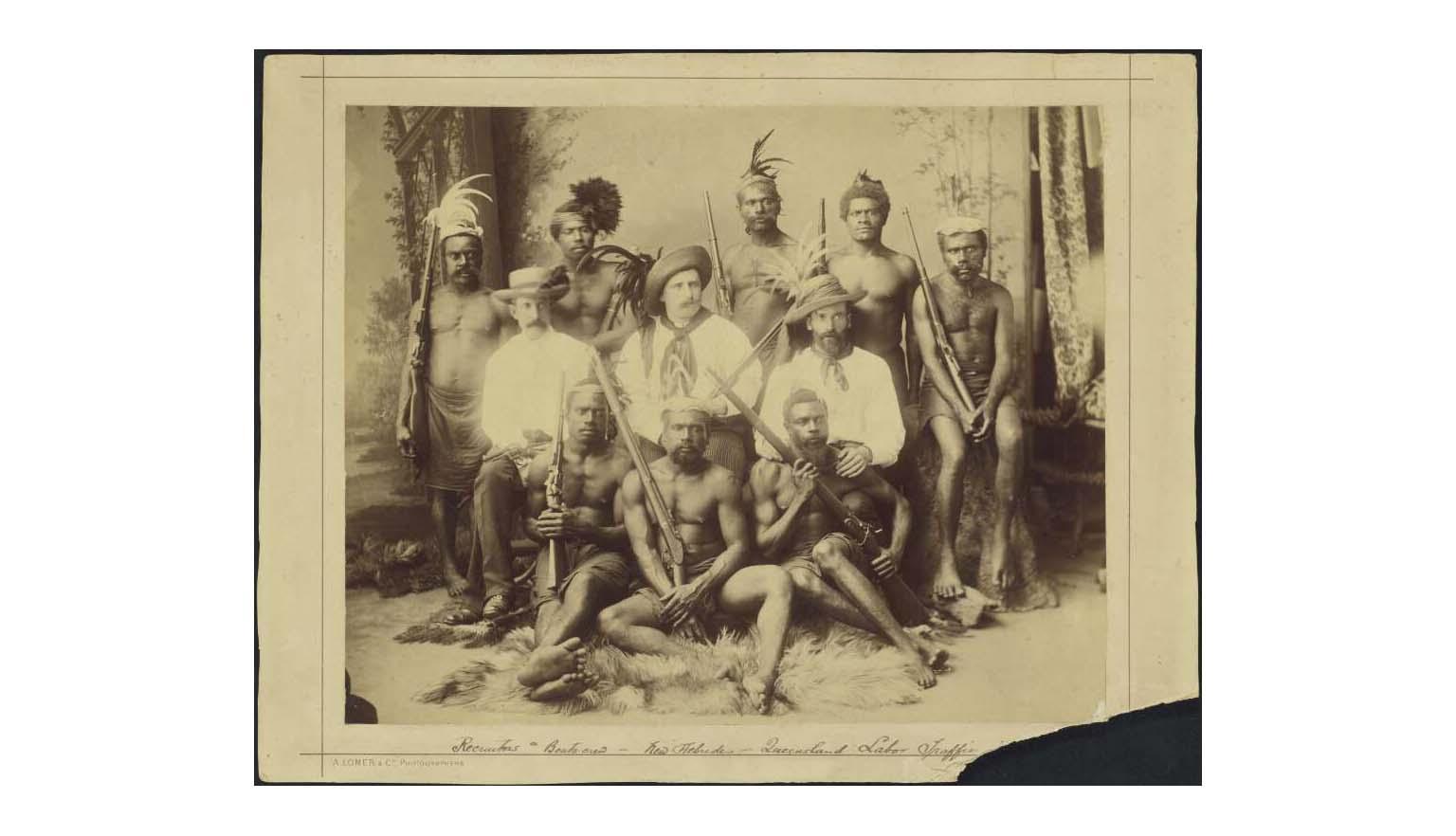 Group portrait of eight Pacific Islander men holding large guns and three British 'recruiters'