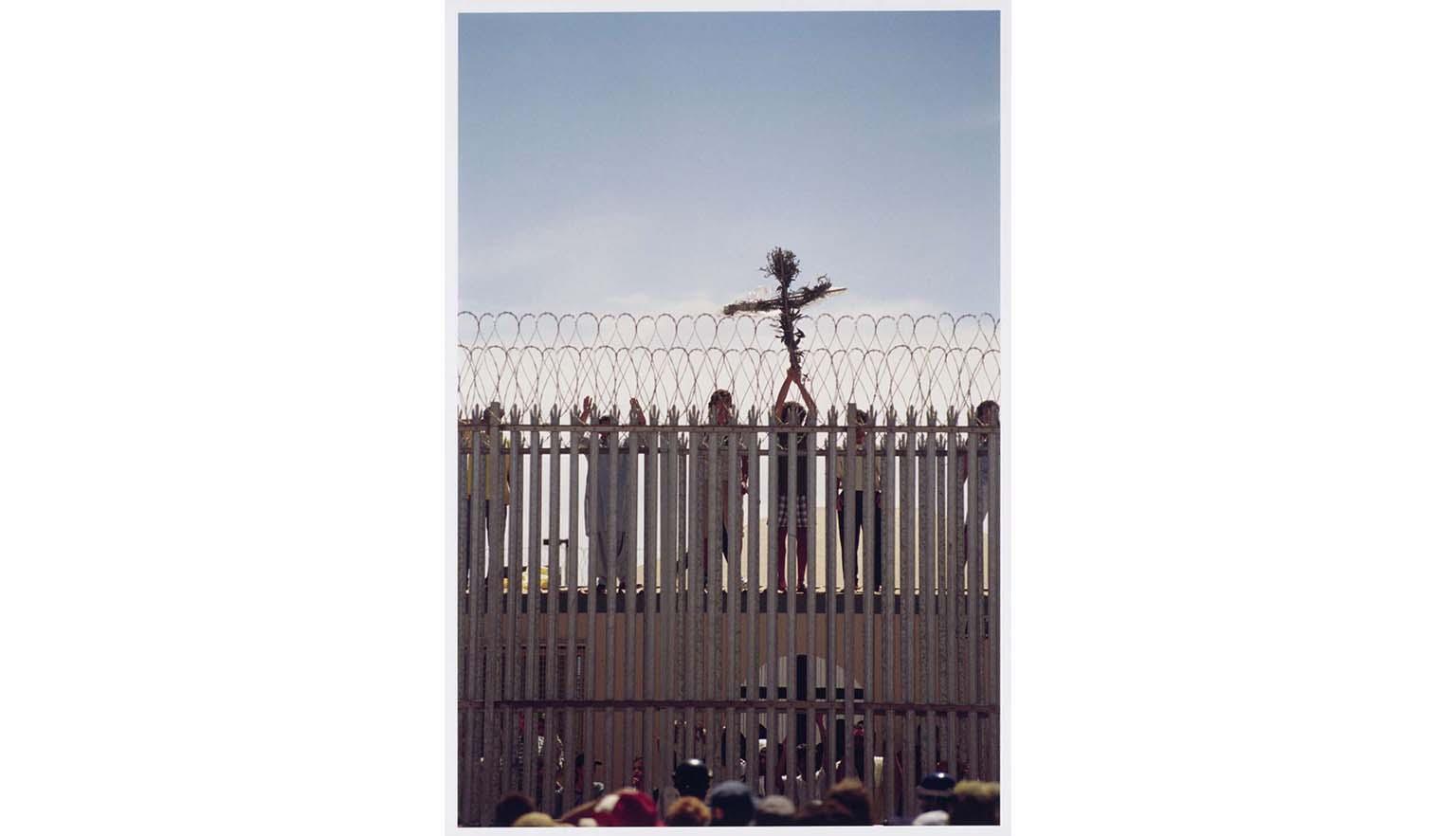 People standing behind a tall fence topped with barbed wire, one of whom is holding up a cross