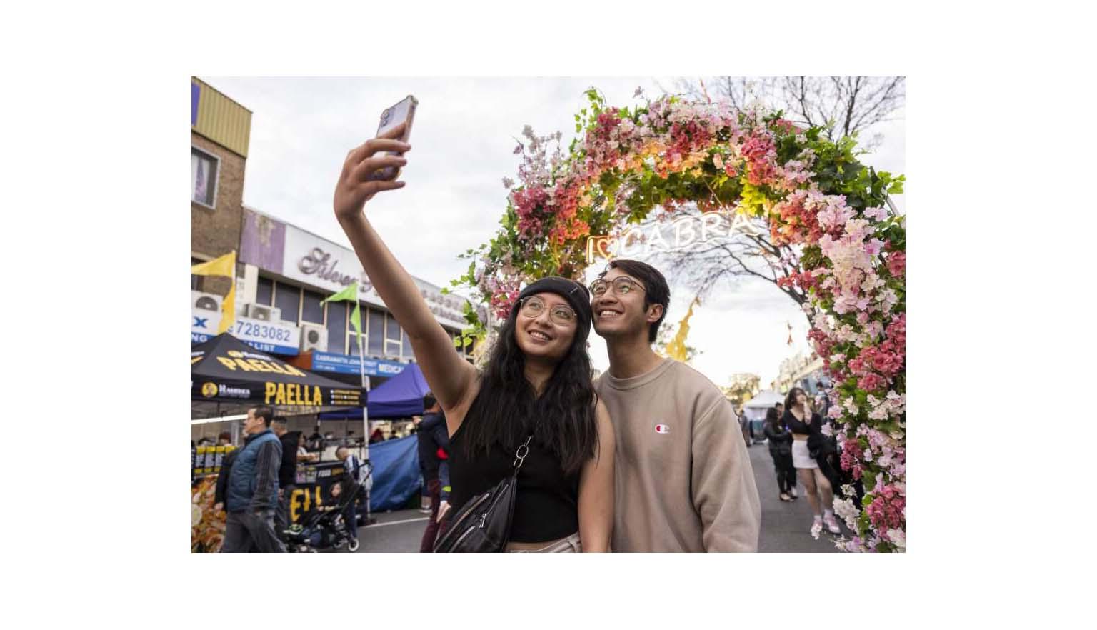 Young man and woman smiling and posing for a selfie in front of a large flower arch with a neon sign reading 'I heart CABRA'