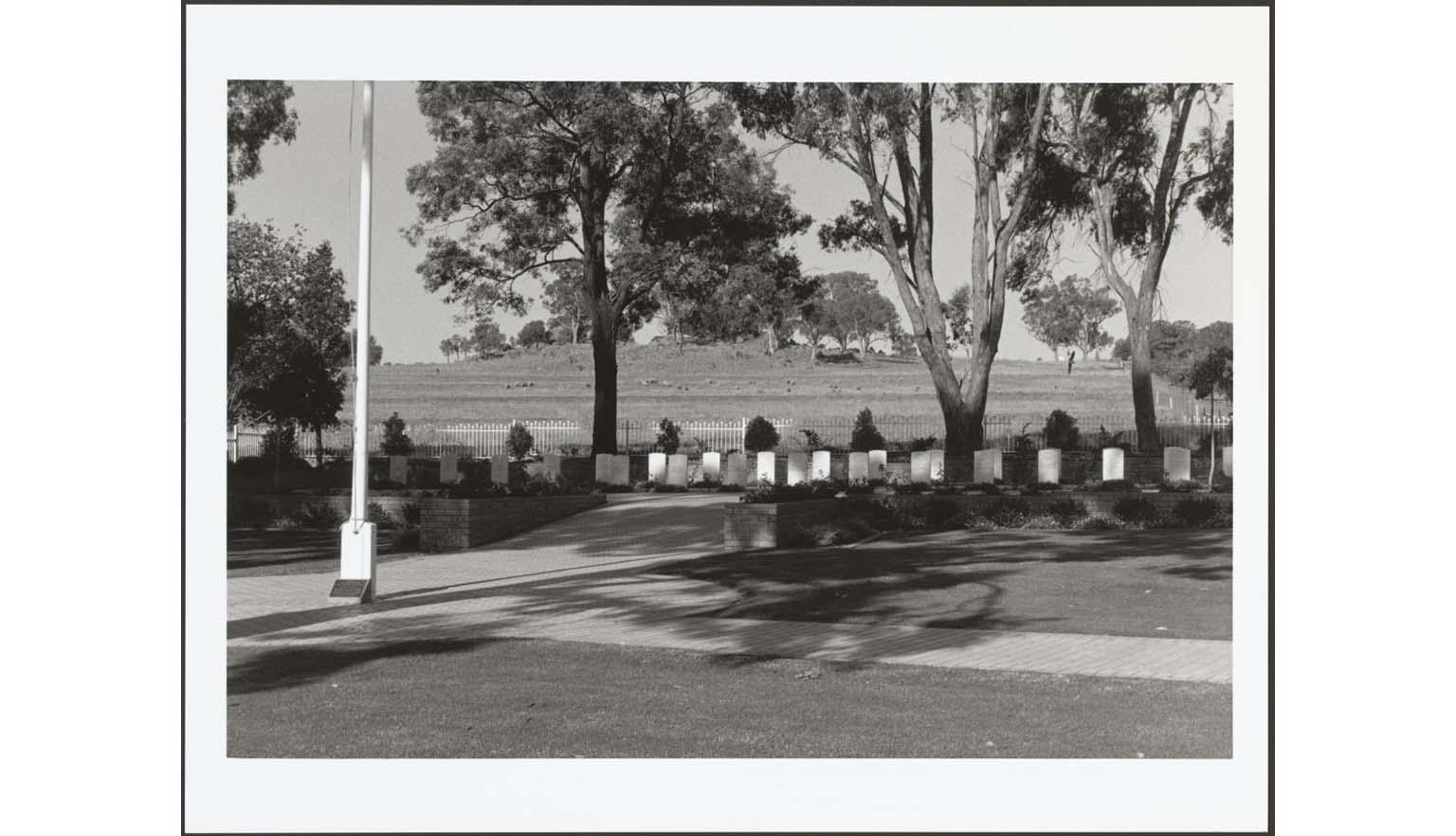 A black and white photograph of a memorial area. There are a row of 23 white headstones along the middle of the image. They are in the shade of large gumtrees. There is a white flagpole on the left of the image. In the background there is a grassy hill.