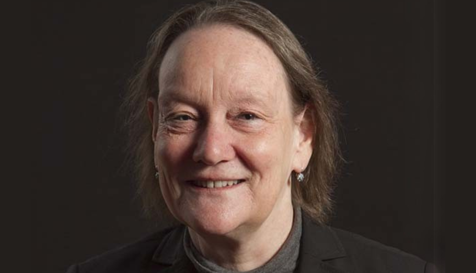 A portrait of a smiling woman wearing a black cardigan over a black top and a grey sweater. She is also wearing a colourful brooch shaped like a tree with colourful leaves.