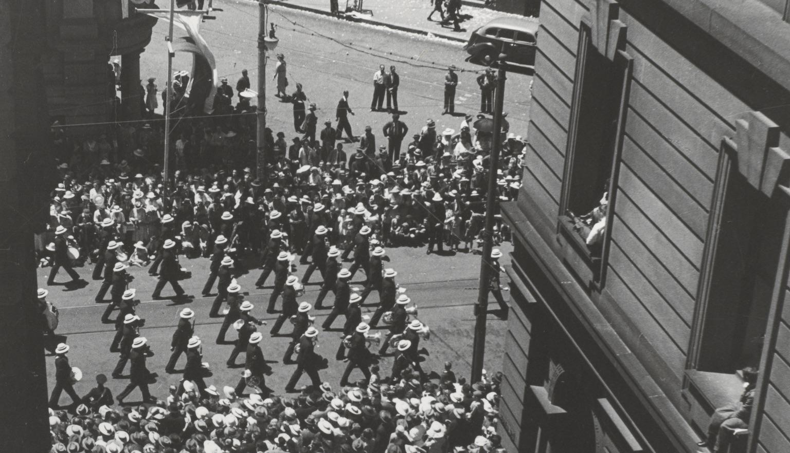 Military procession down a street surrounded by crowds, viewed from above