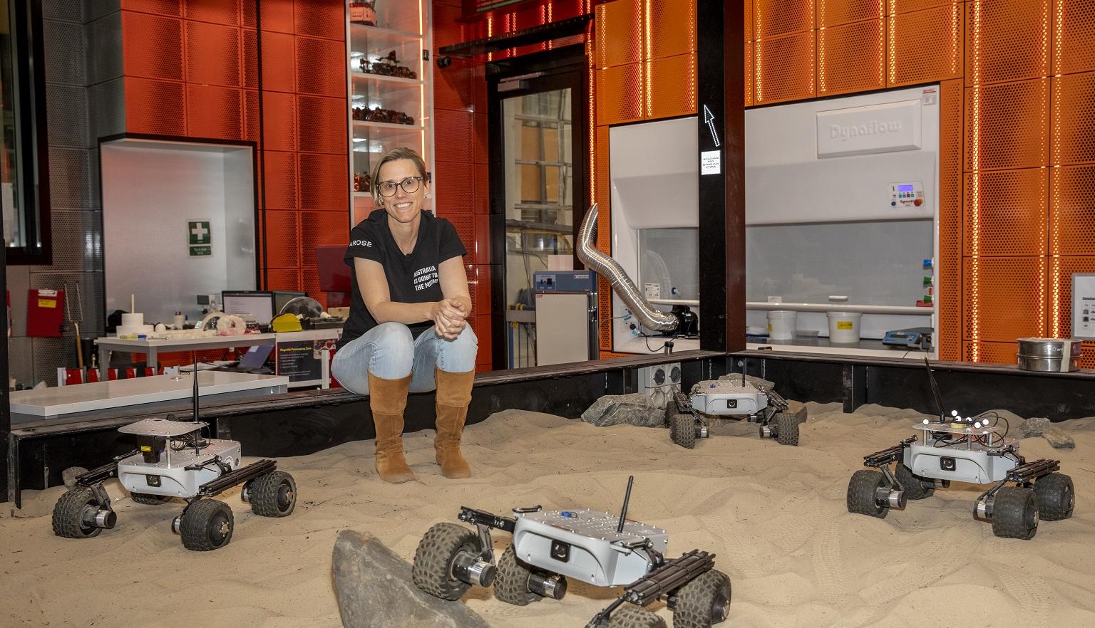 Dr Sarah Cannard sitting on the edge of a sand pit with four space rovers in it