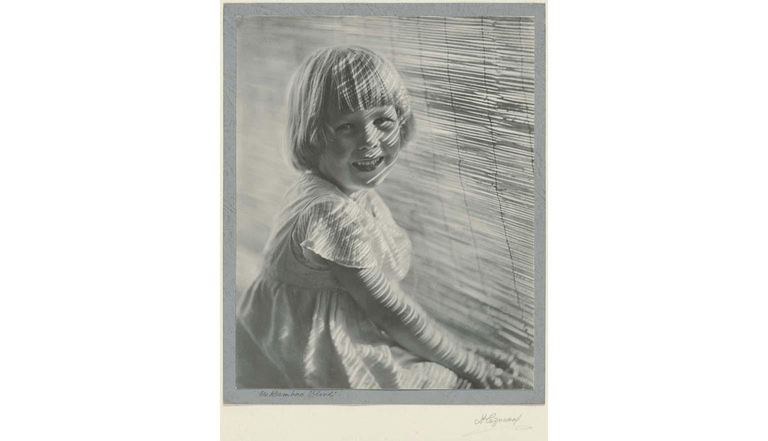 Black and white photo of a young girl smiling at the camera and sitting in front of a bamboo blind with sunlight lines across her face. 'The Bamboo Blind' is handwritten on the bottome left and 'H Cazneeaux' on the bottom right