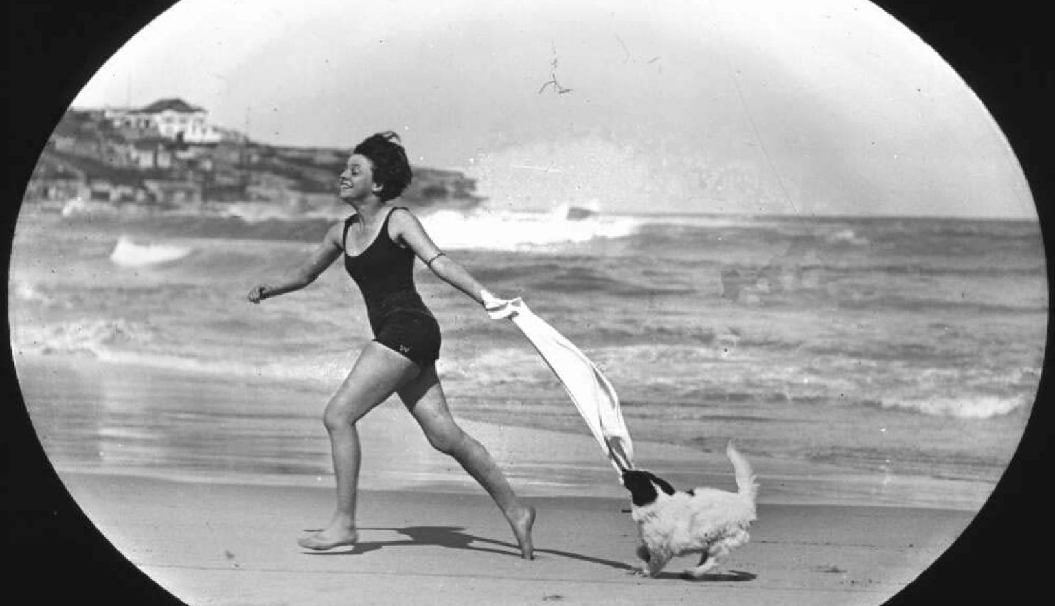 Black and white photo of a woman in a swimsuit and with a towel running on a beach with a small dog running behind her