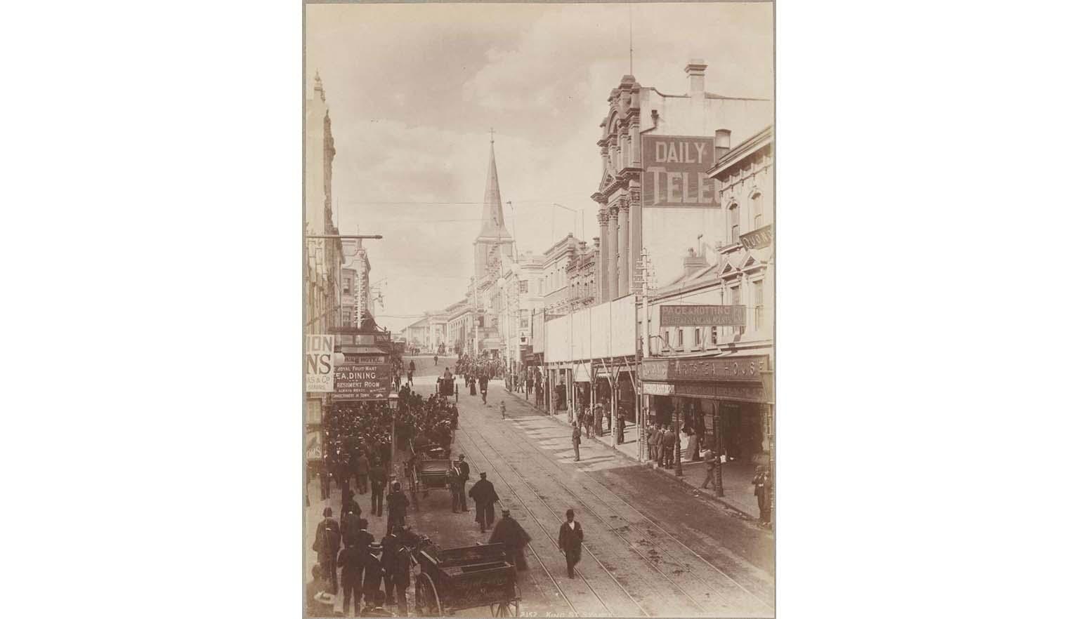 Sepia photo of a dirt street with buildings on either side, people standing and walking, and horses and carts