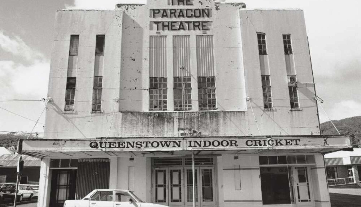 Black and white photo of a whit building with the words 'The Paragon Theatre' at the top and an old car parked at the front under an awning
