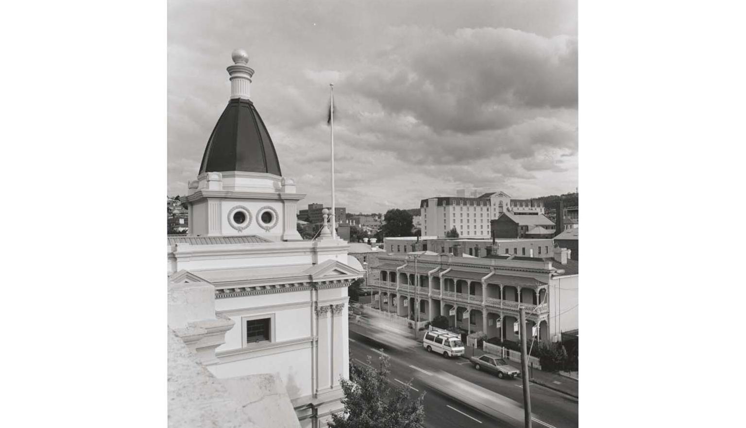 Black and white photo looking down from a roof, with a cupola on the building on the left and terrace houses across the street with cars parked outside