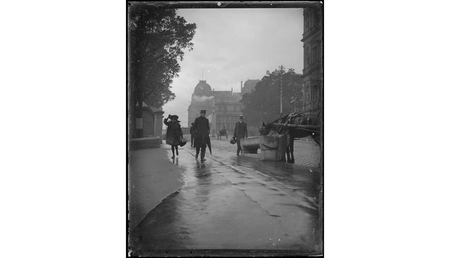 Black and white photo of a wet street showing buildings, people walking with hats and umbrellas, a horse drinking from a trough, and a horse and cart in the background