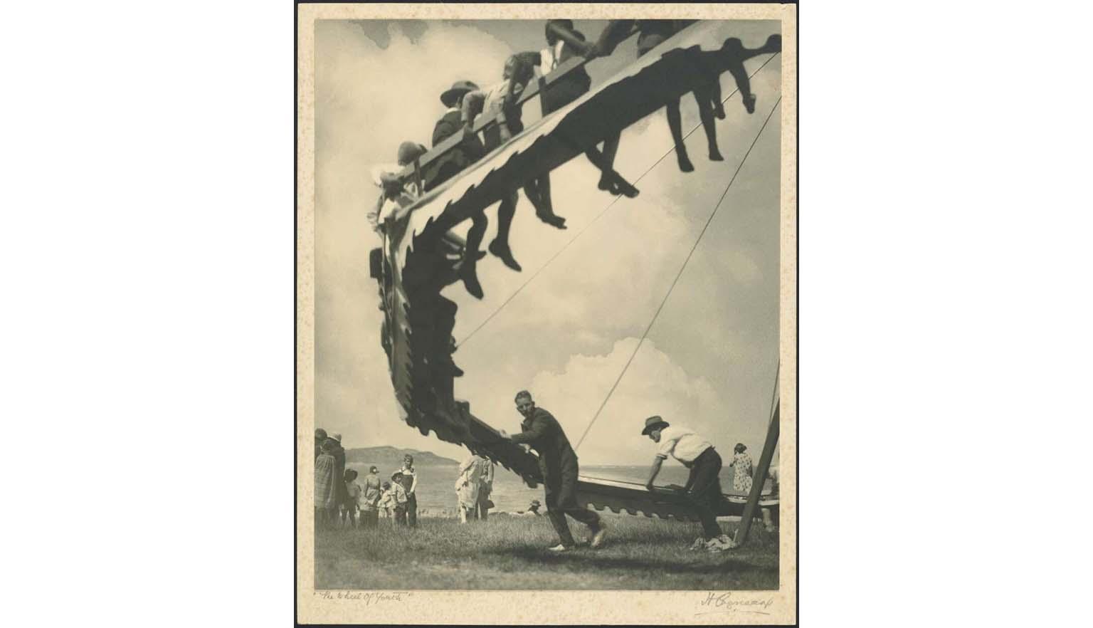 Sepia-toned photograph of people being swung on a round wheel. 'The Wheel of Youth' is handwritten on the bottom left and 'H Cazneeaux' on the bottom right