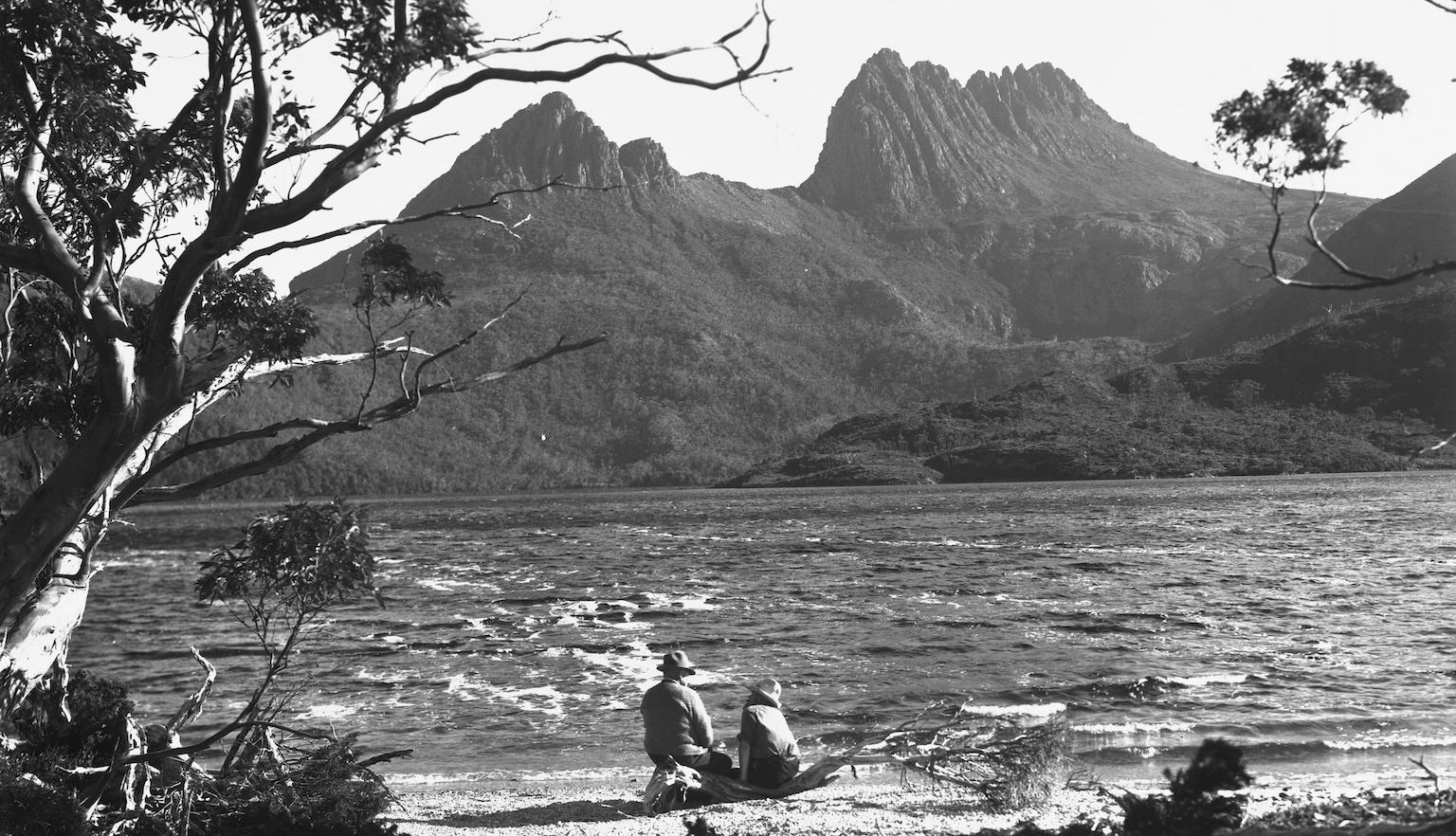 Two people sit on the bank of Dove Lake looking out to Cradle Mountain