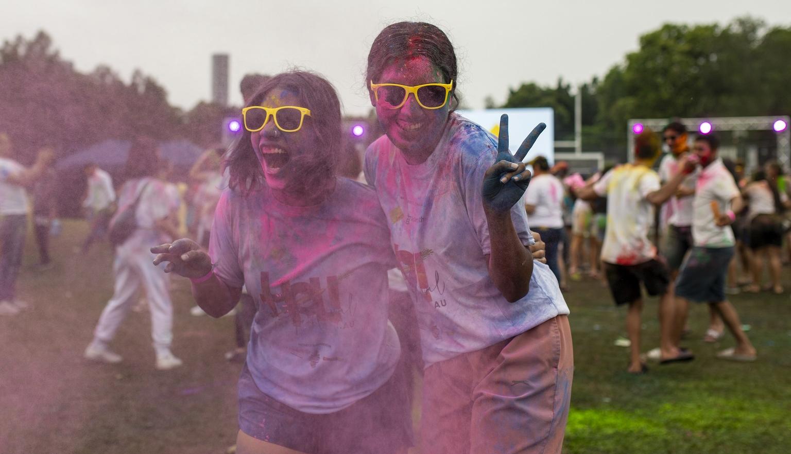 Two young girls posing with peace signs, covered in multi-coloured powder gulal at Holi Festival in a park