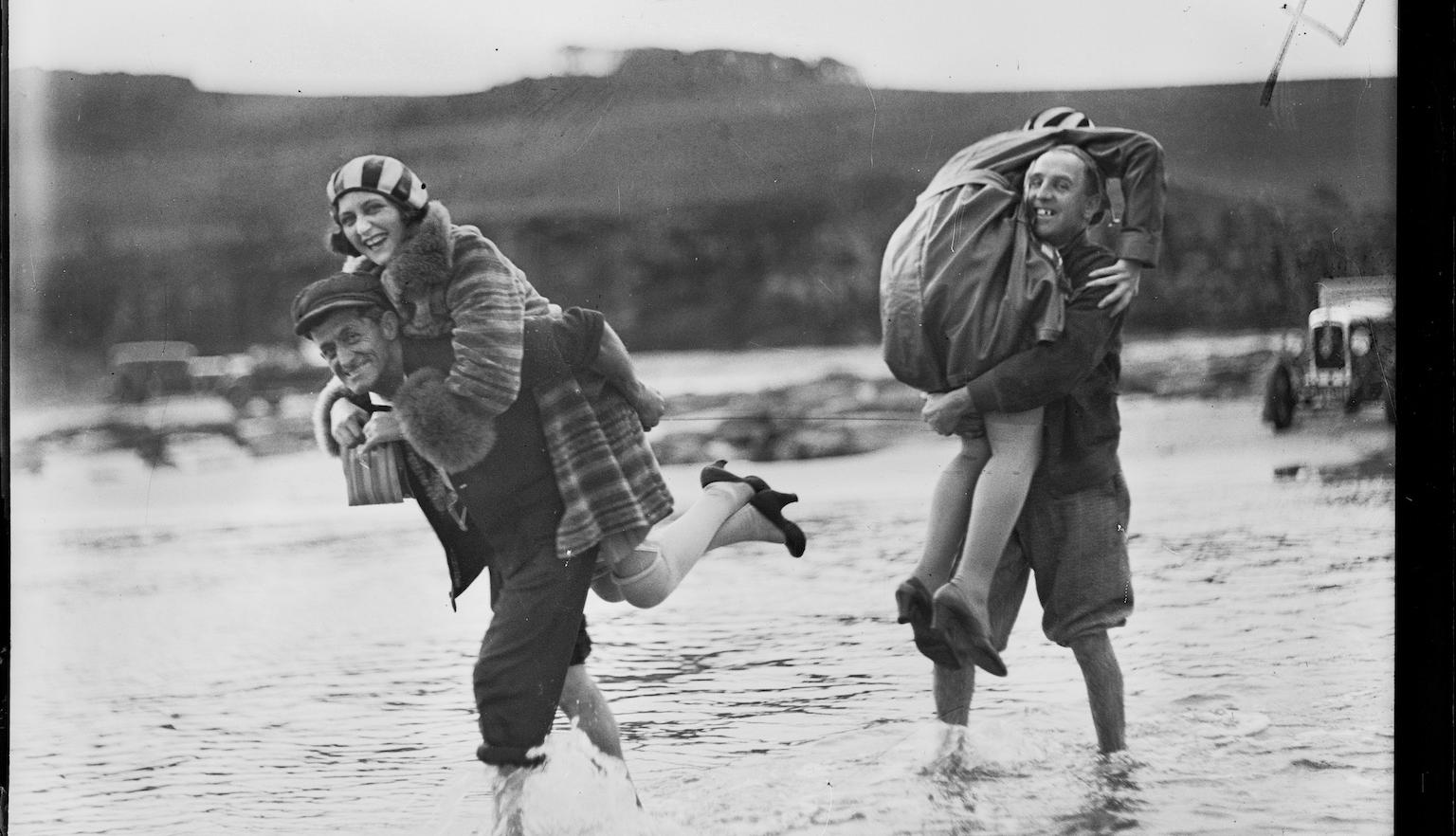 Black and white photo of two women laughing while being carried by two men over a flooded racetrack