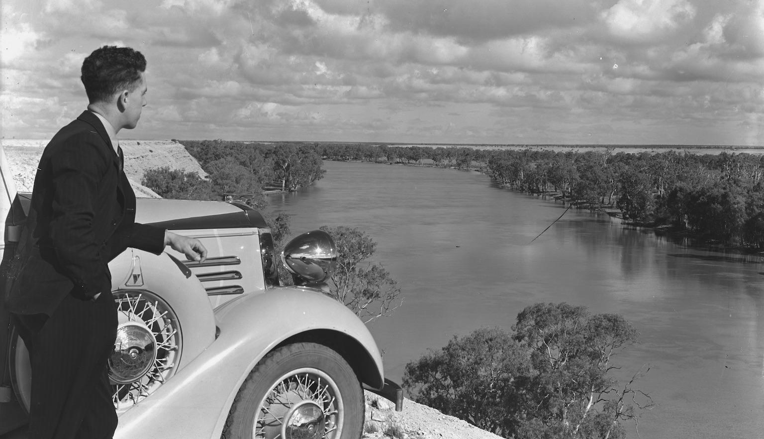 A man dressed in a suit standing beside the front of car on a clifftop overlooking the tree lined Murray River.  