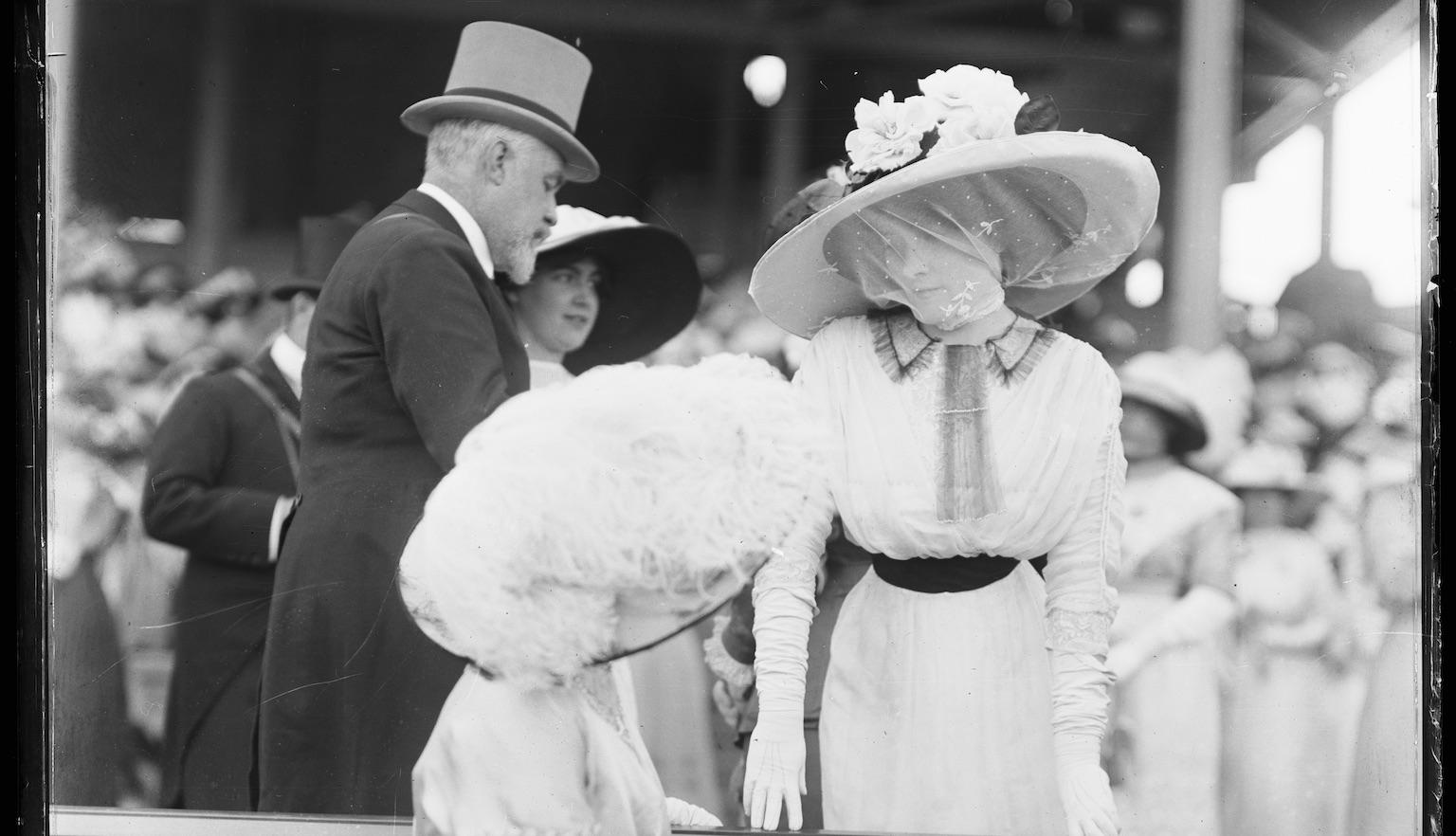 Black and white photo of two women in fancy dresses and hats chatting near a bench