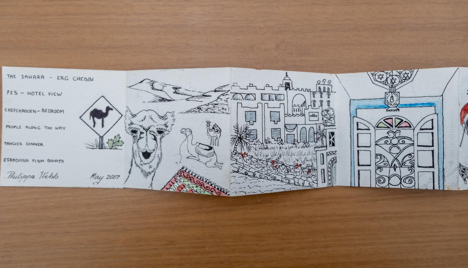 Fold-out travel diary open to three pages with black and white illustrations of camels, Moroccan buildings and a door