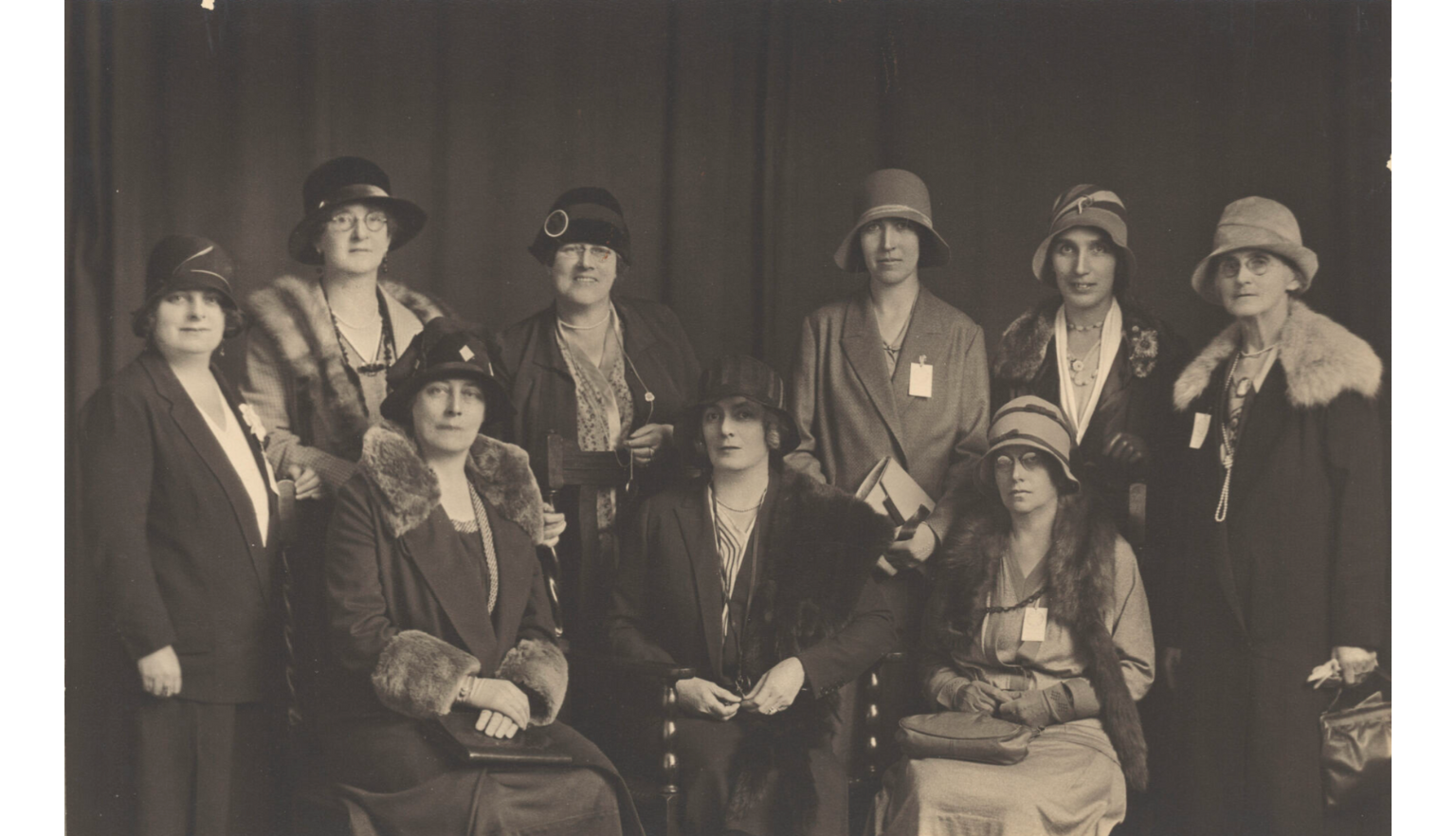 Nine members of the Australian Federation of Women Voters posing for a group photo, some standing and some sitting, in two rows in front of some dark curtains