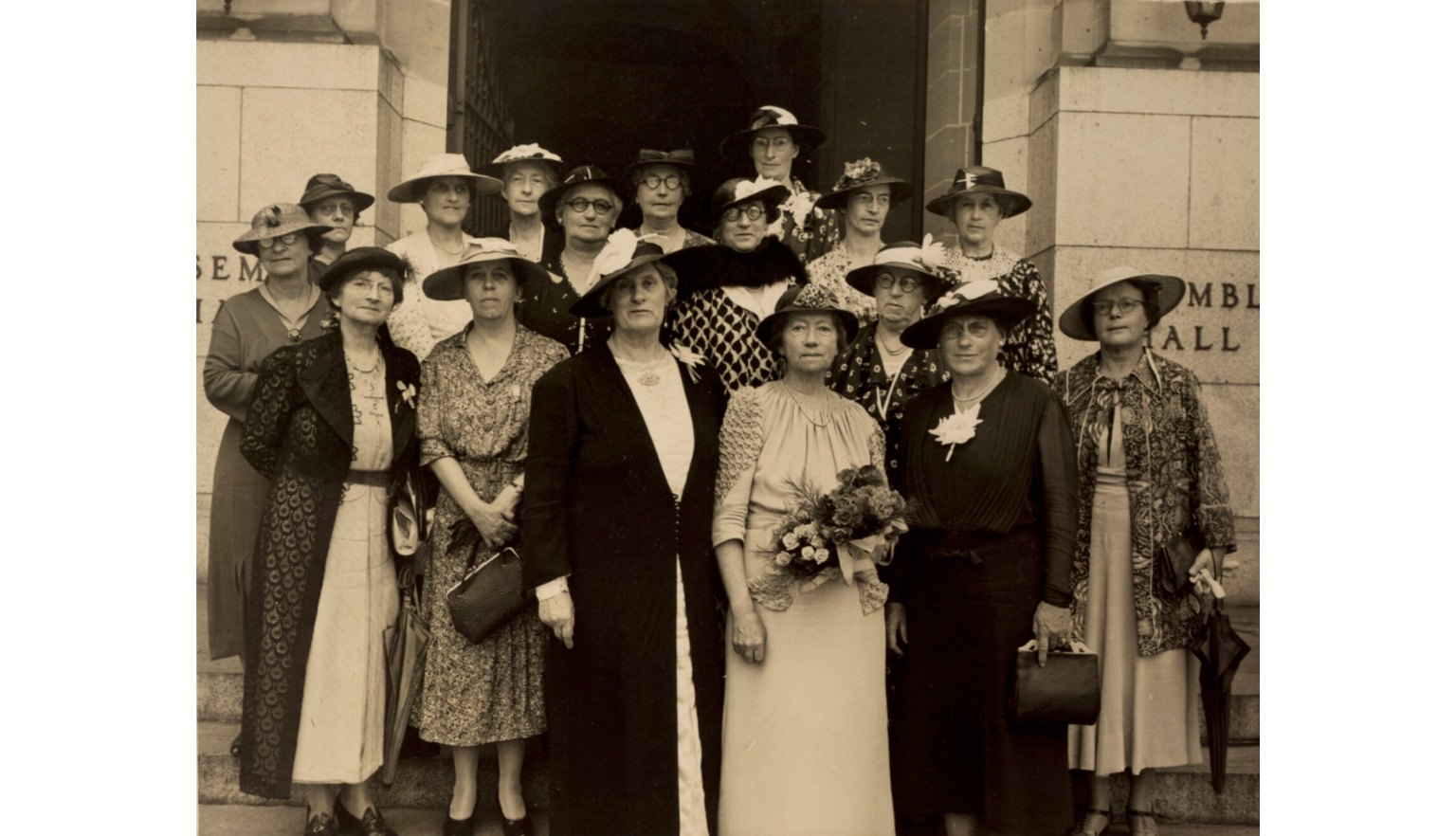 Sepia-toned photo of 16 members of the Australian National Council of Women standing with Dame Maria Ogilvie Gordon on the steps in front of a building
