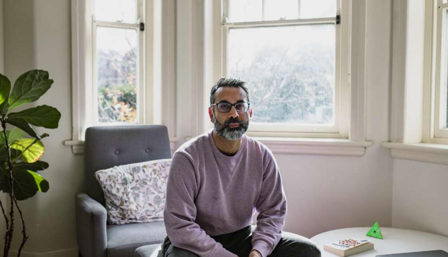 A man wearing glasses and a purple jumper siting in a room