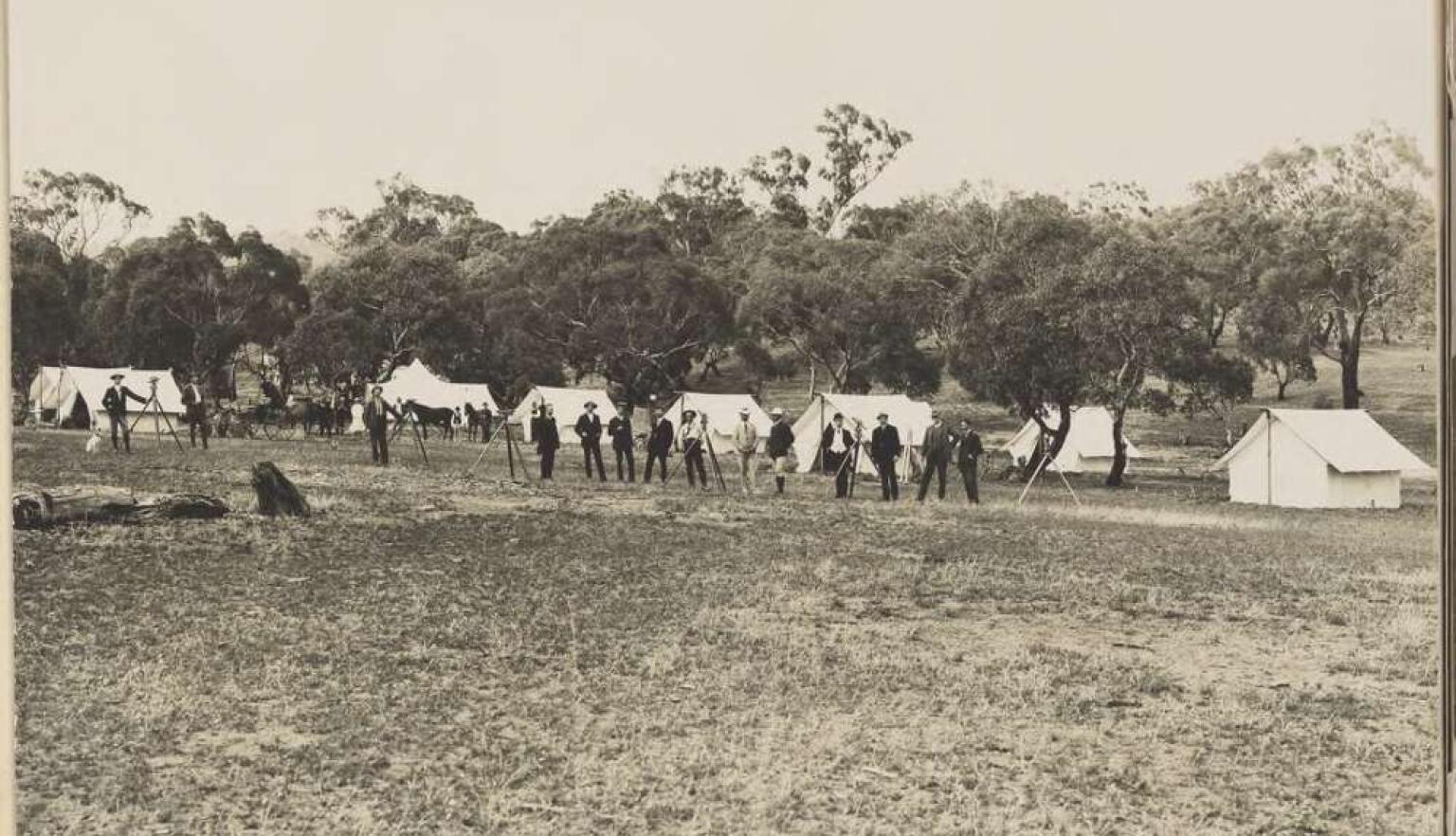 A black and white image of men surveying land, with tents in the near background