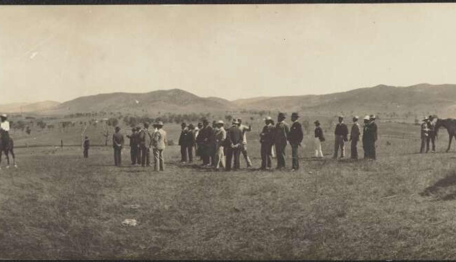 A black and wihte photo of a group of men standing in a field, some on horseback, some on carts, inspecting the field