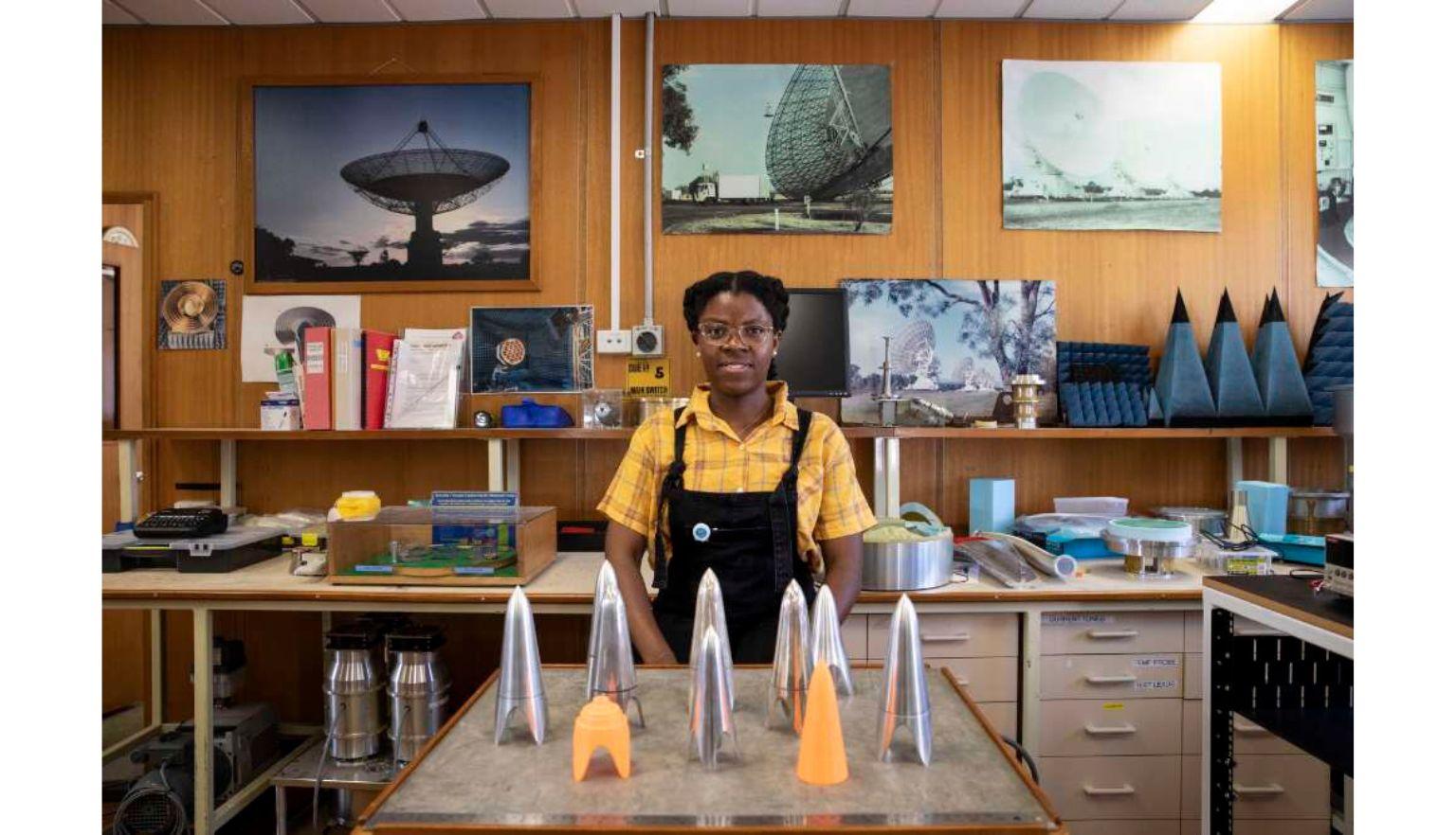 Natasha Maimbo, a young woman of colour, standing in a mechanical engineering lab with pictures of telescopes behind her.