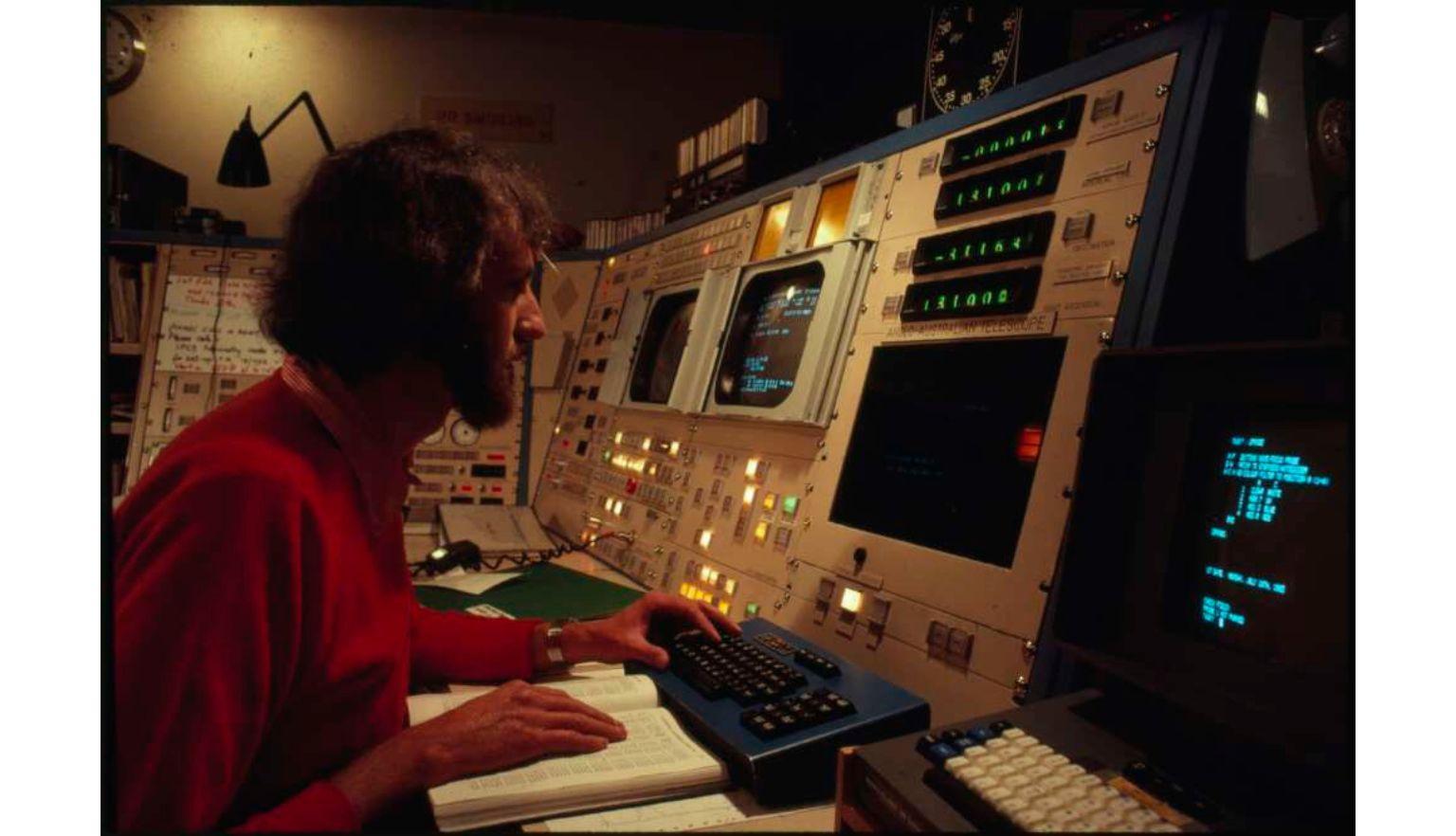 A photograph of a man sitting at a control panel comprised of computer screens, lights, and switches.
