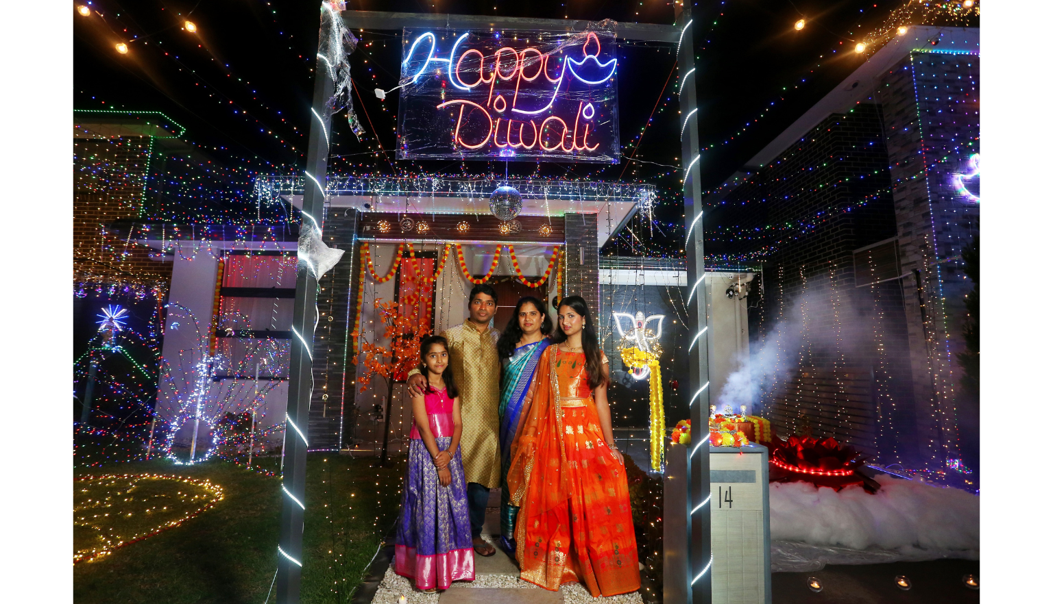 Family standing in front of a house covered in colourful lights, with one reading 'Happy Diwali' at night