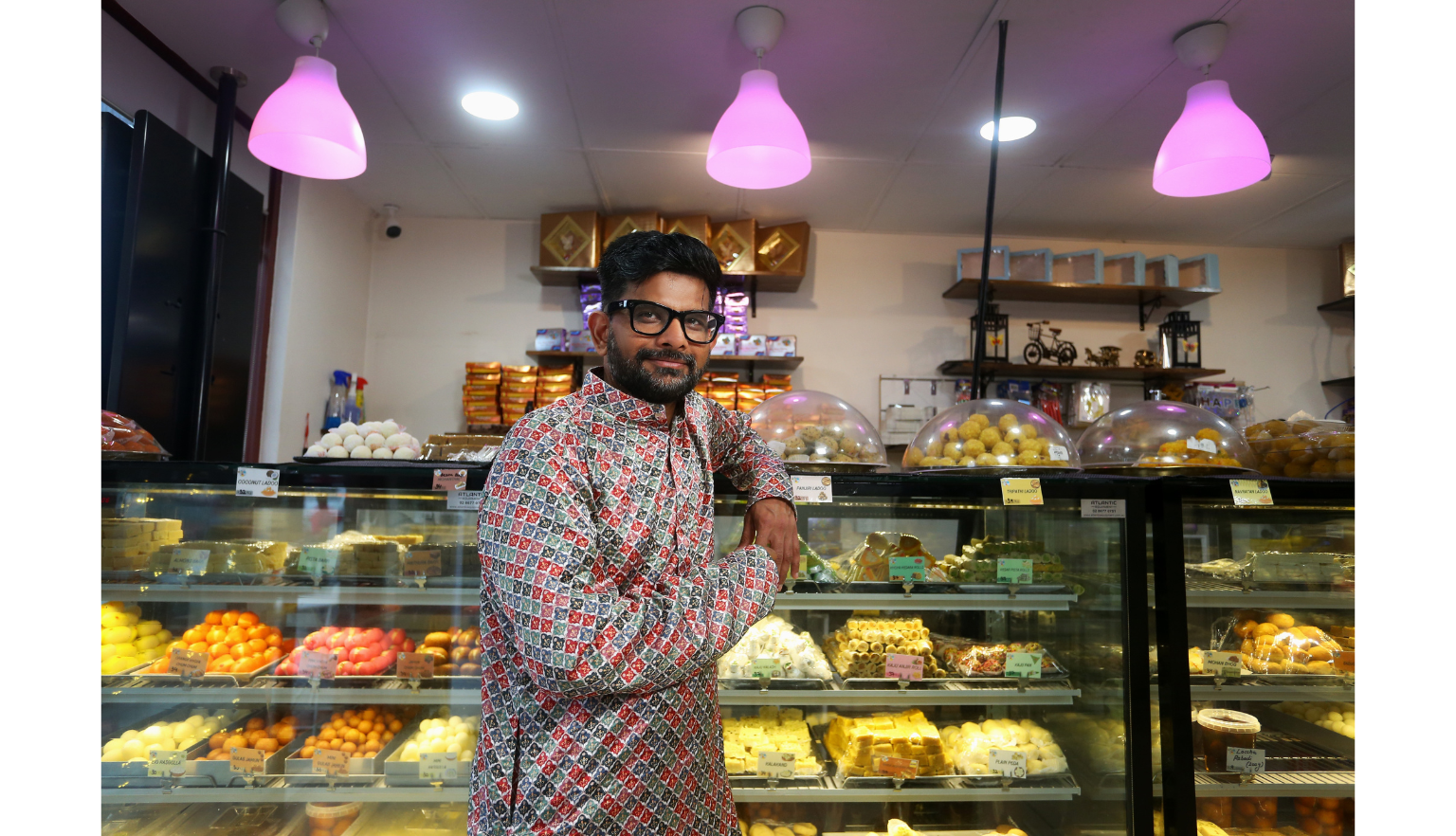 Man standing and leaning on a display case of traditional Indian sweets