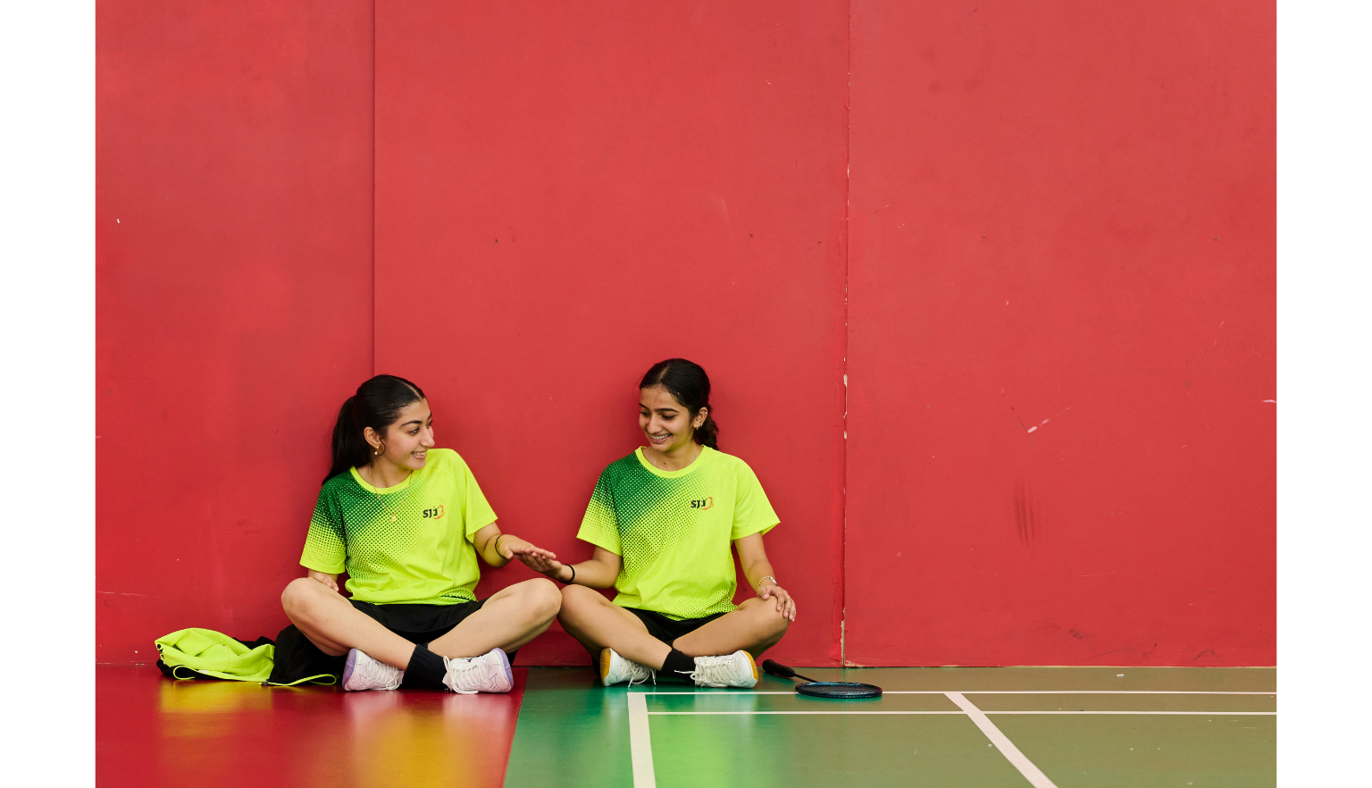 Two girls in bright yellow and black sports uniforms sitting against a red wall