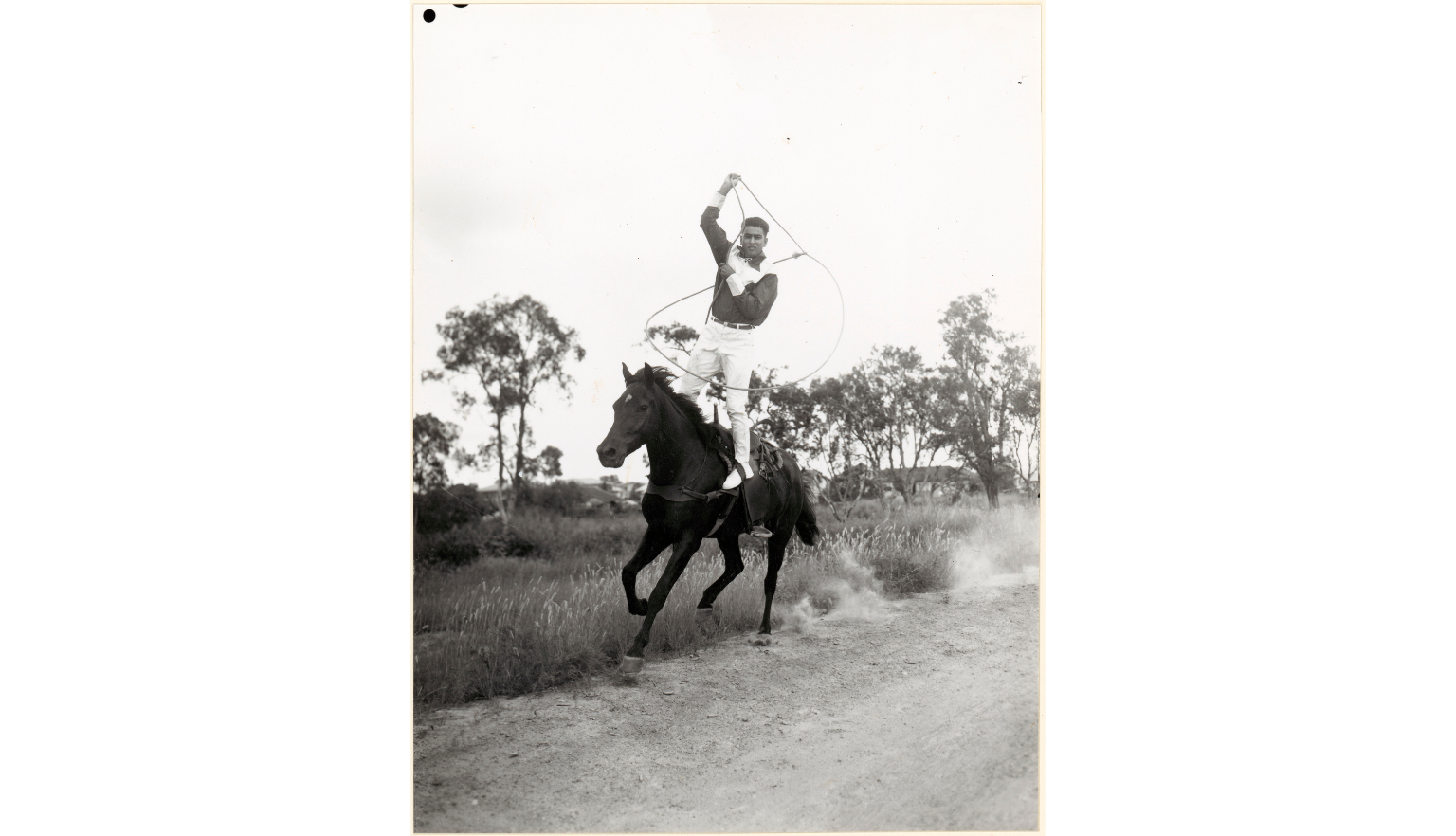 Black and white photo of a standing on the back of a horse swining a lasso above his head