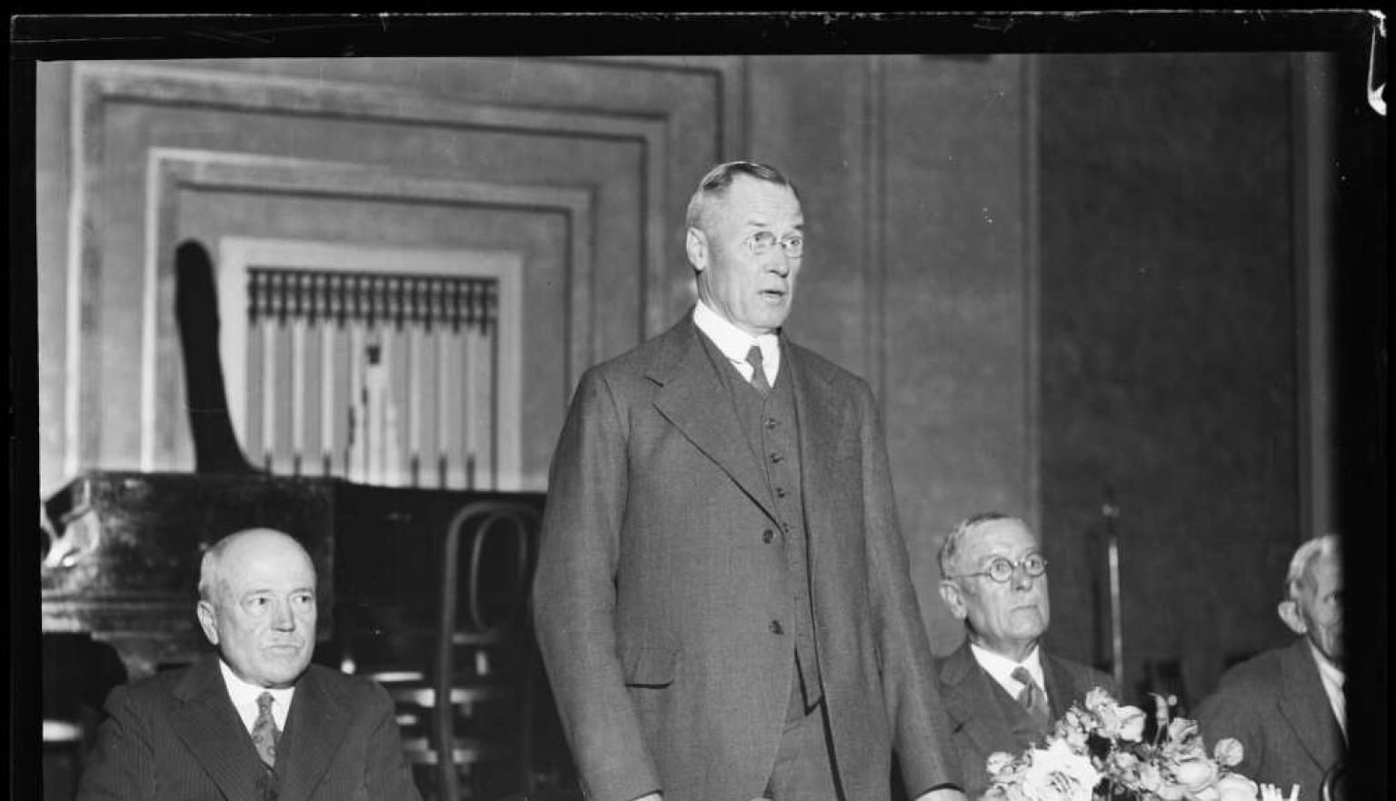 A gentleman standing behind a table in a suit addressing the room 