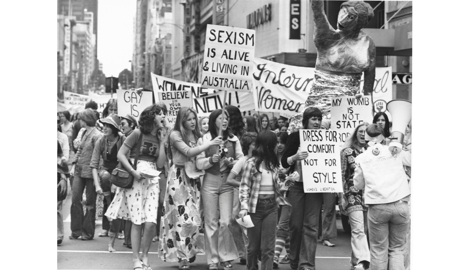 A group of women marches down a city street, holding various protest signs. Signs include messages like "Sexism is alive & living in Australia" and "Dress for comfort, not for style." A large sculpture is visible in the background.