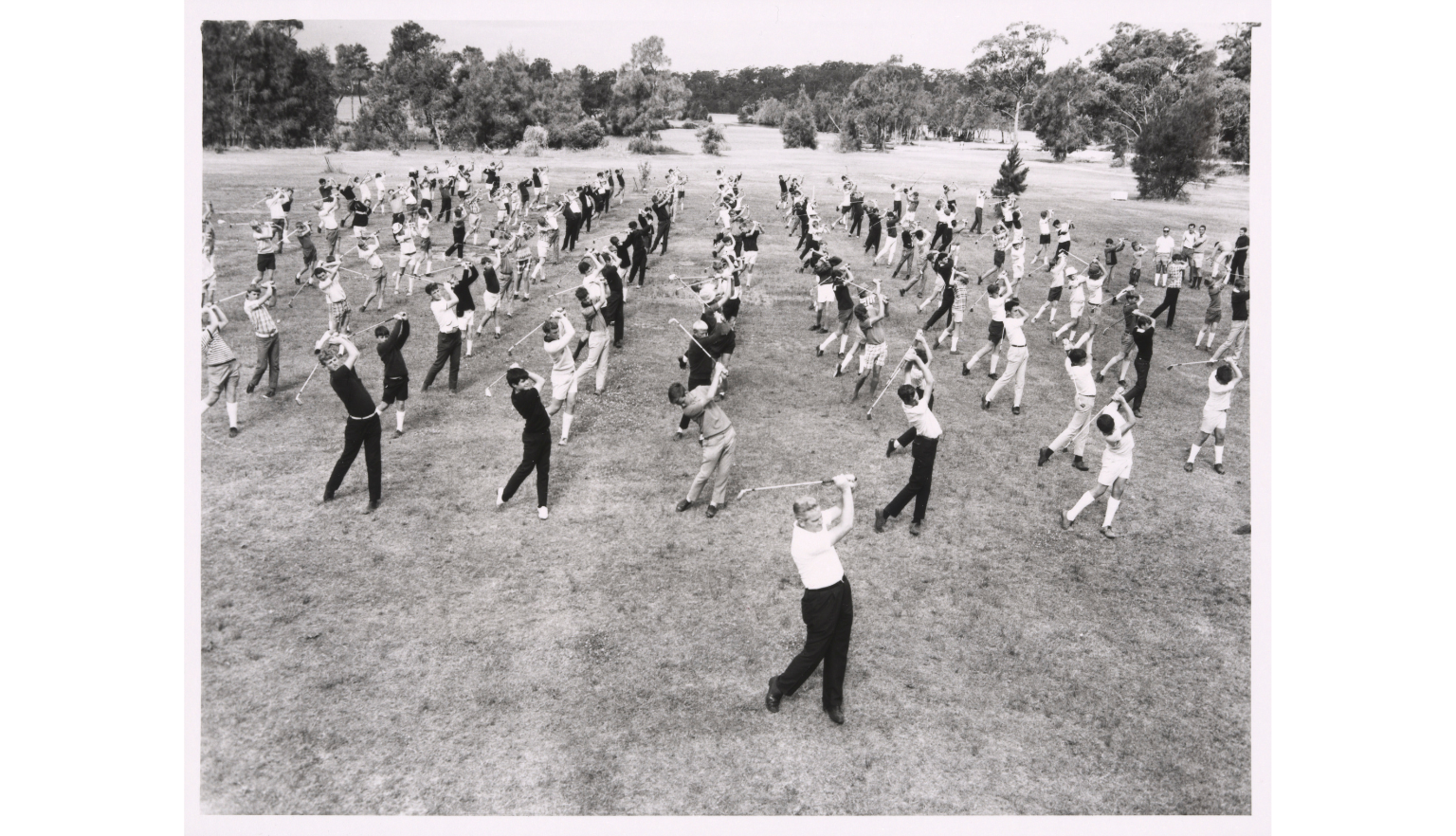 A black and white photograph showing a large group of people practicing a golf swing