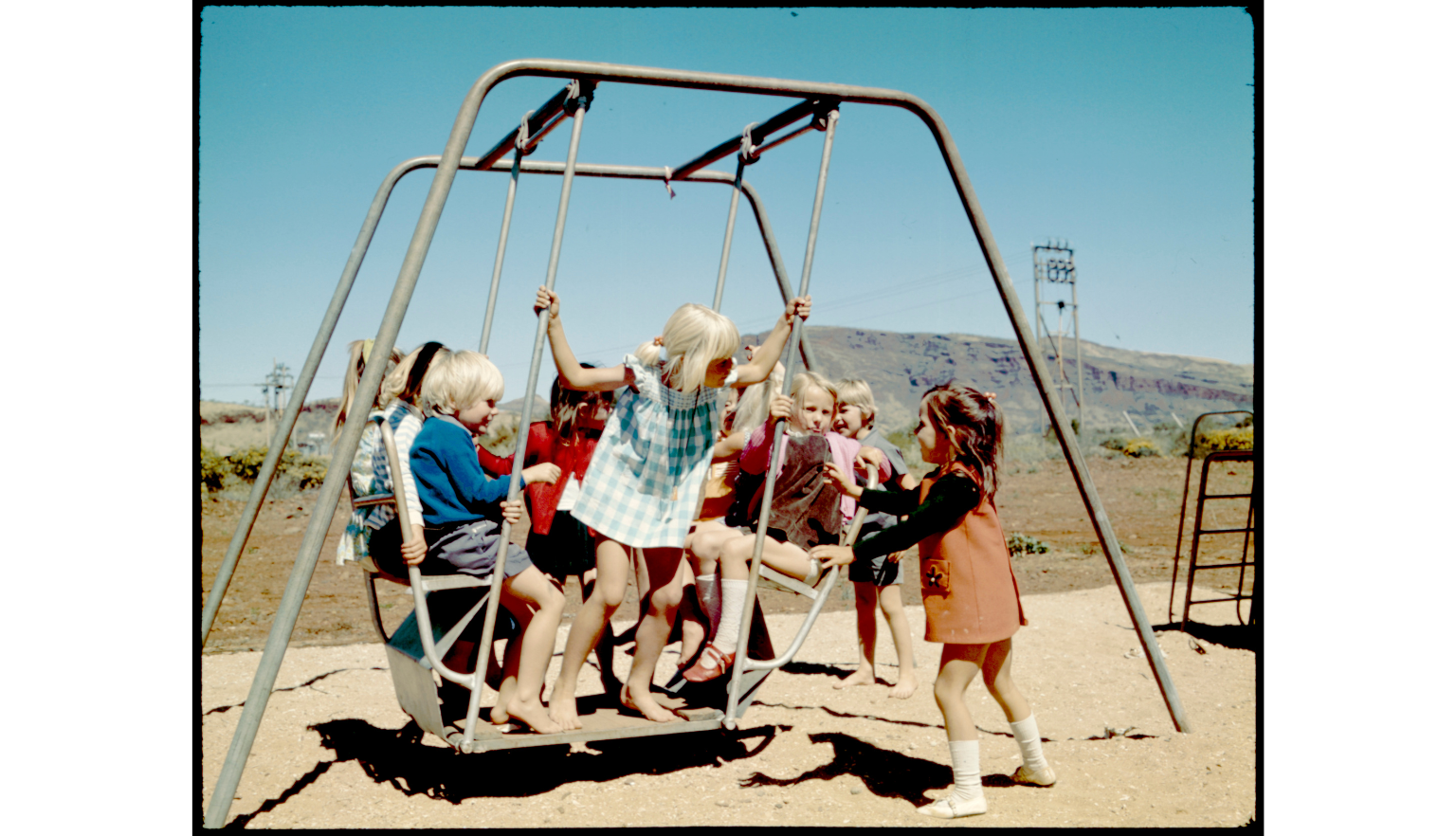 Photo of several young children in 1970s-era clothing playing on a swing set on a sunny cloudless day