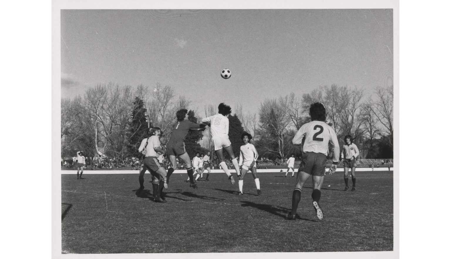 A black and white photograph showing a soccer match. Some players are jumping to contest a header 