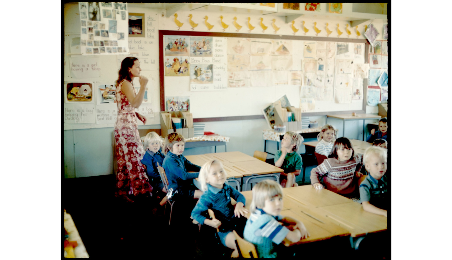 A colour photograph showing a teacher and children in a classroom in the 1970s