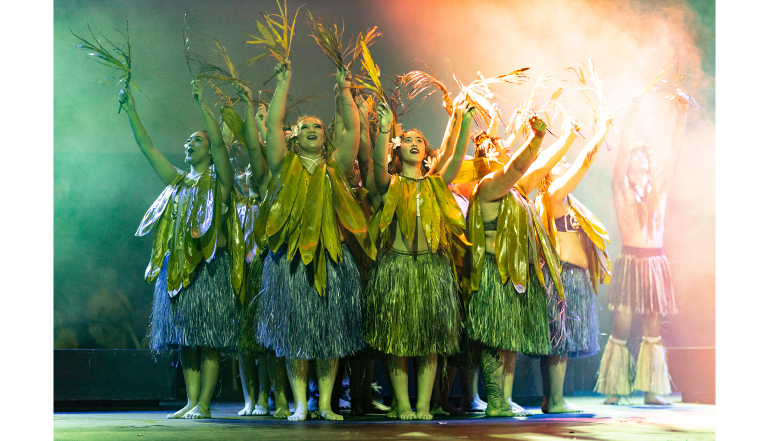 Group of dancers wearing large leaves around their necks and grass skirts, standing in a circle with their arms raised