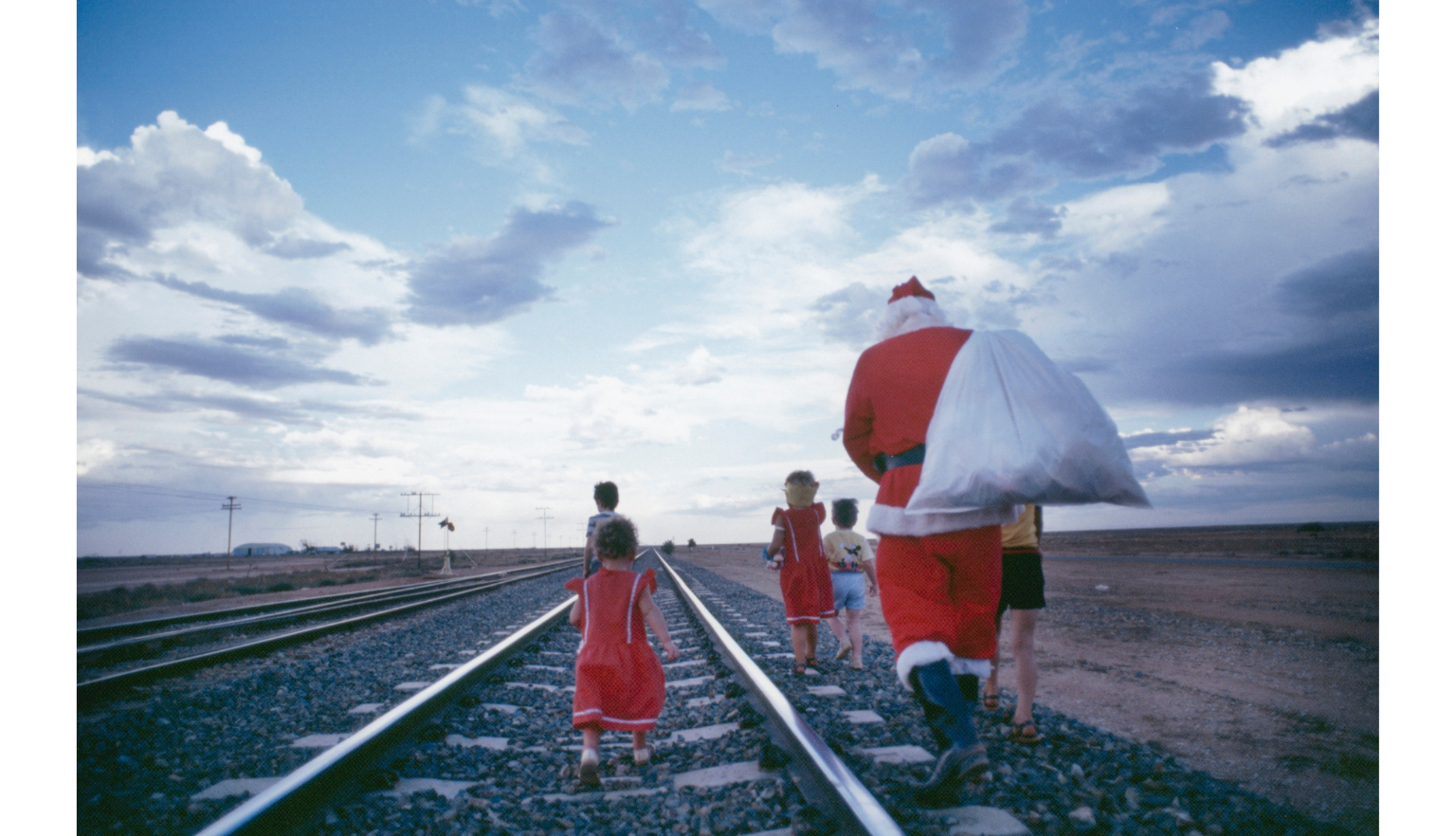 Man in a Santa suit carrying a large sack over his shoulder walking along a train track with 5 children
