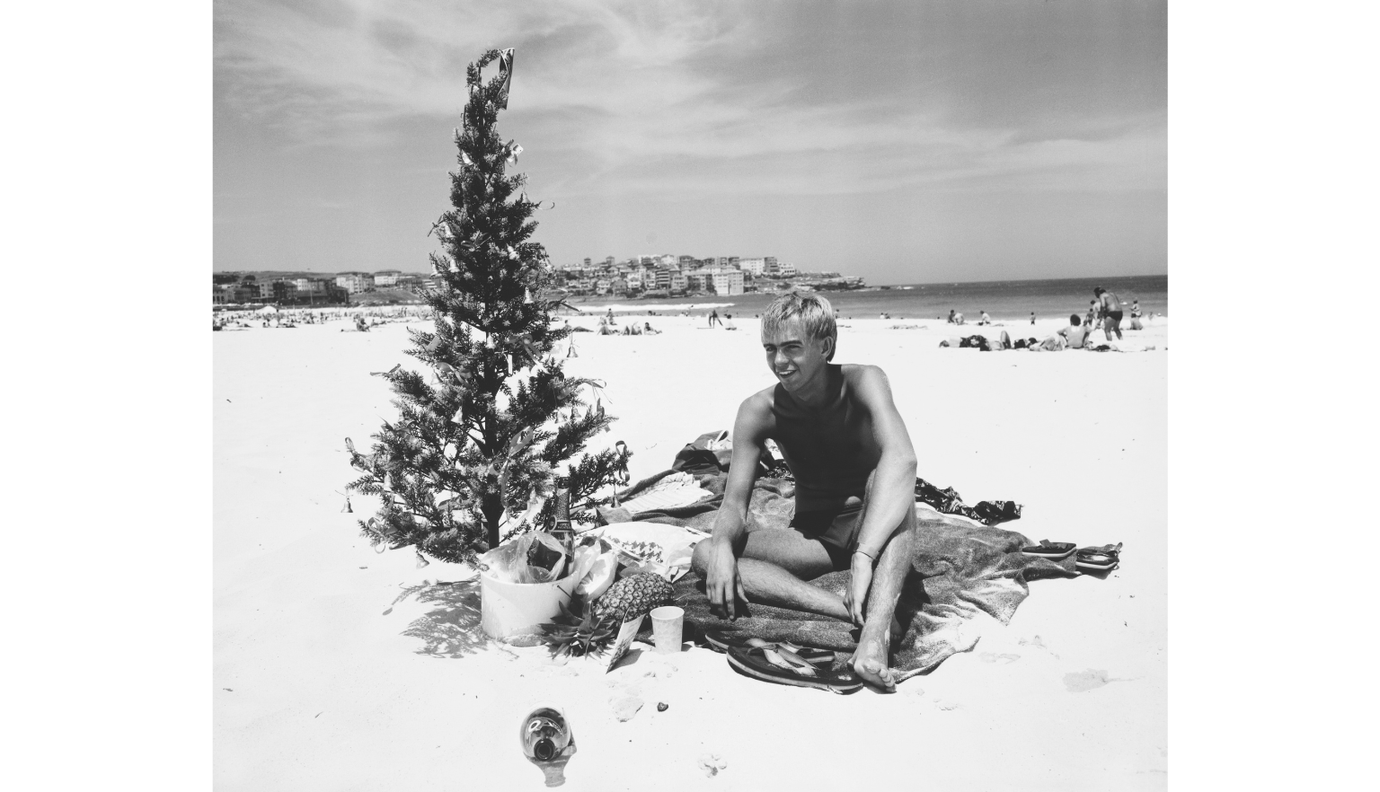 Black and white photo of a man sitting on a beach next to a Christmas tree