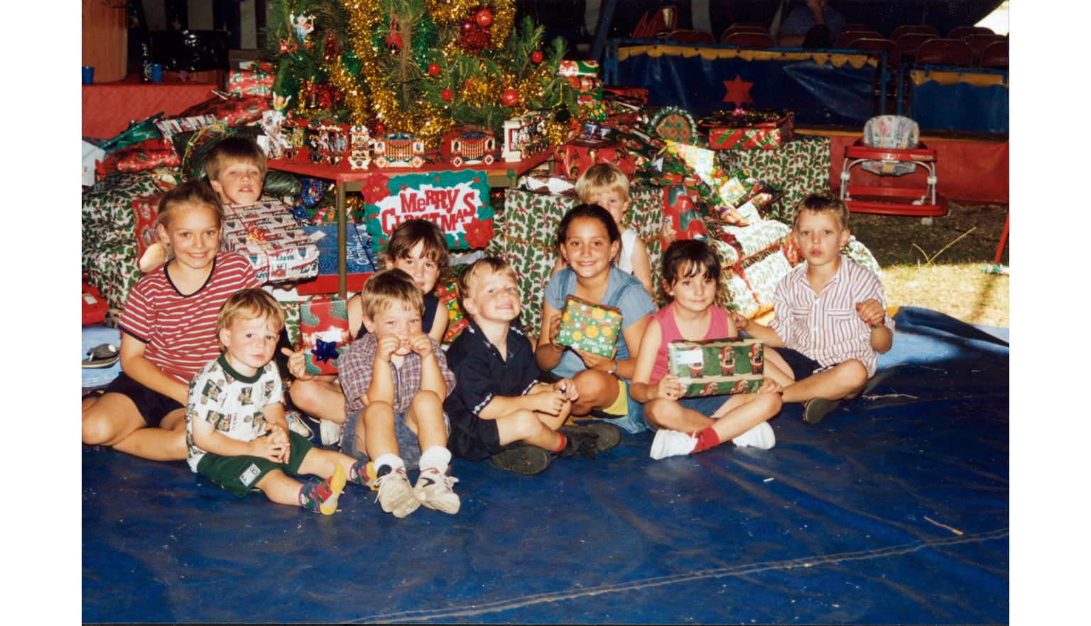Group of 10 young children sitting under a Christmas tree with presents
