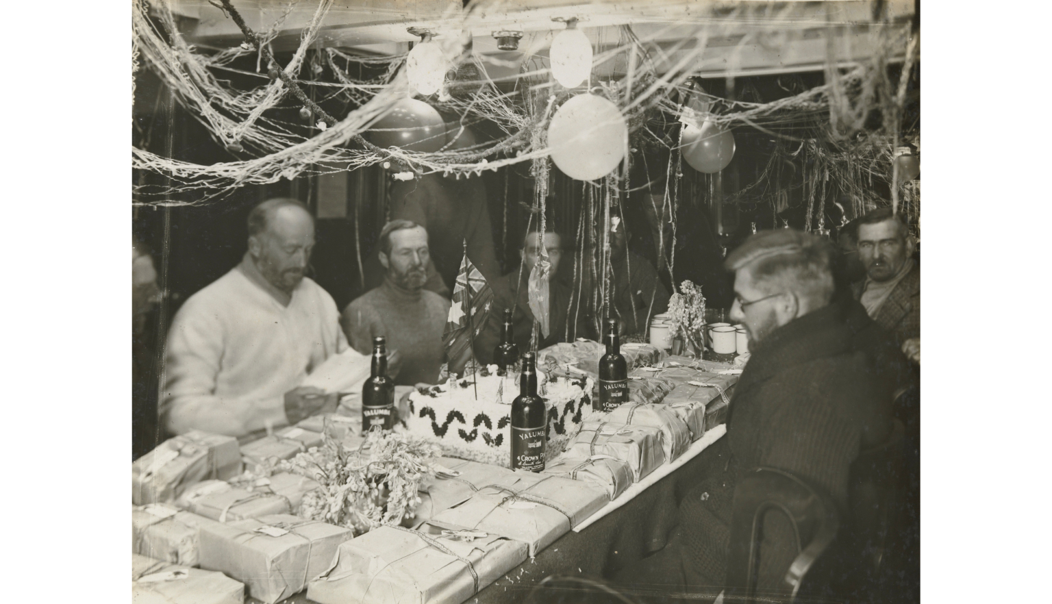 Black and white photo of men sitting at a table covered in small packages in a room decorated with streamers and balloons