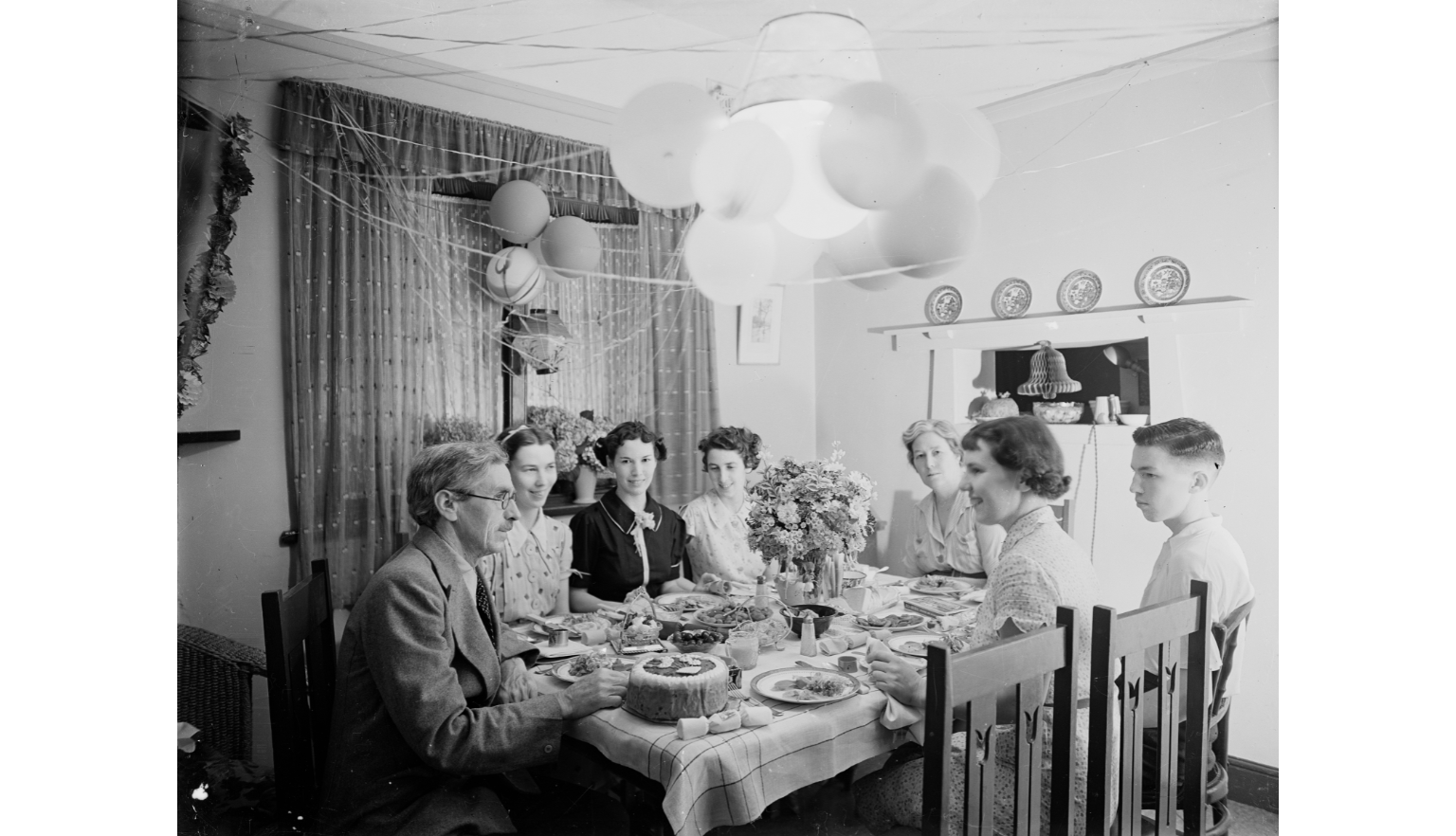 Black and white photo of a family sitting around a table, decorated with streamers and balloons, for Christmas dinner