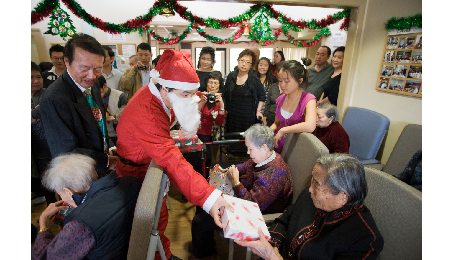 Man in a Santa costume handing out presents to residents of Elderly Chinese Home, in a room decorated with red and green tinsel
